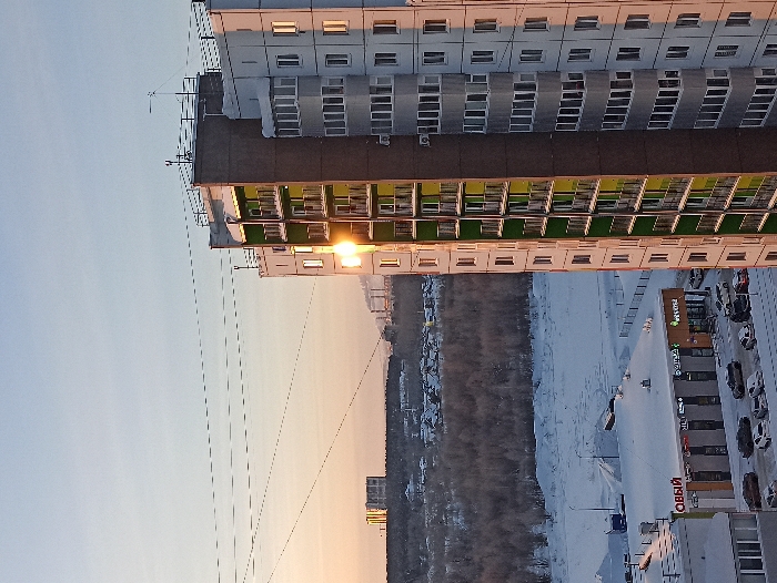 A high-angle view shows a tall building with rows of windows, some reflecting sunlight. Adjacent is a street lined with shops and parked cars, covered with snow. A dense forest stretches beyond, also blanketed in snow. The sky is light, with a gradient of blue and orange, suggesting sunrise or sunset. Power lines are visible across the sky.
