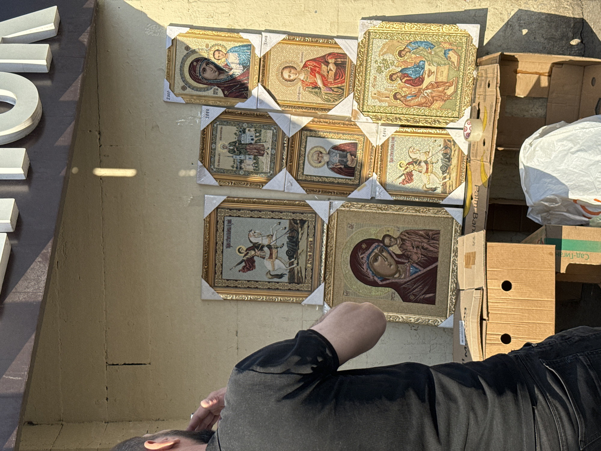 The image shows a display of framed religious icons on a pale wall outdoors, next to cardboard boxes. A man in a black shirt is visible in the foreground, apparently looking at the icons. The icons are in gold frames, with various religious depictions, some featuring the Virgin Mary and saints. The location is likely in Odintsovo, Russia, based on the Cyrillic text visible on one of the boxes. The scene suggests a street vendor selling religious items.