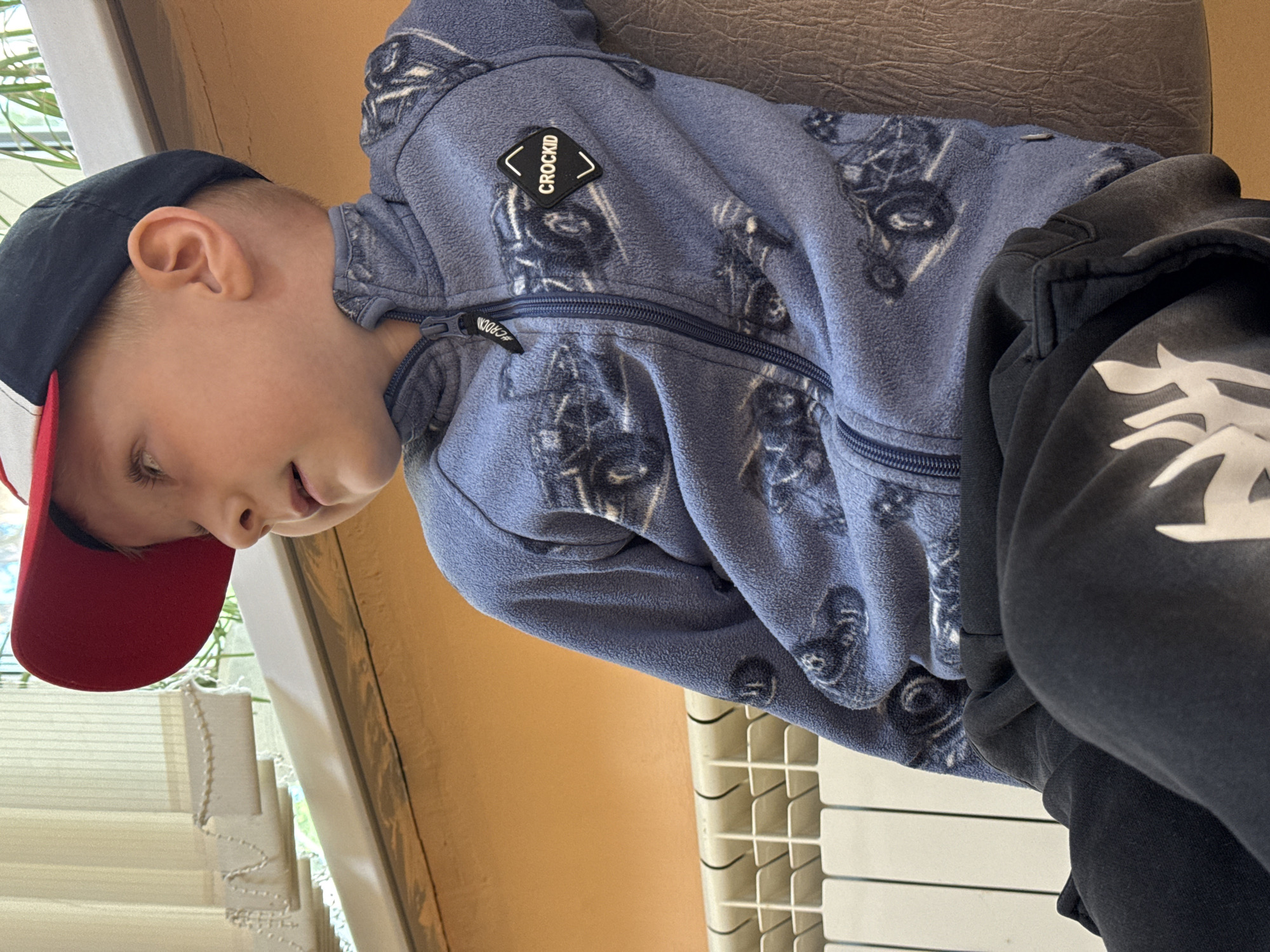 A young boy in a blue patterned fleece jacket and a black and red baseball cap sits in what appears to be an indoor setting. Part of his face is visible, and he seems to be looking down or to the side. The jacket has a logo on the left chest that reads "CROCKID". He is wearing dark pants with white graphics on the right leg. The setting includes a window with blinds and a wall painted in a muted orange color. Part of a white radiator is visible in the background. It is difficult to determine the specific location in Chaikovsky, Russia without additional context.