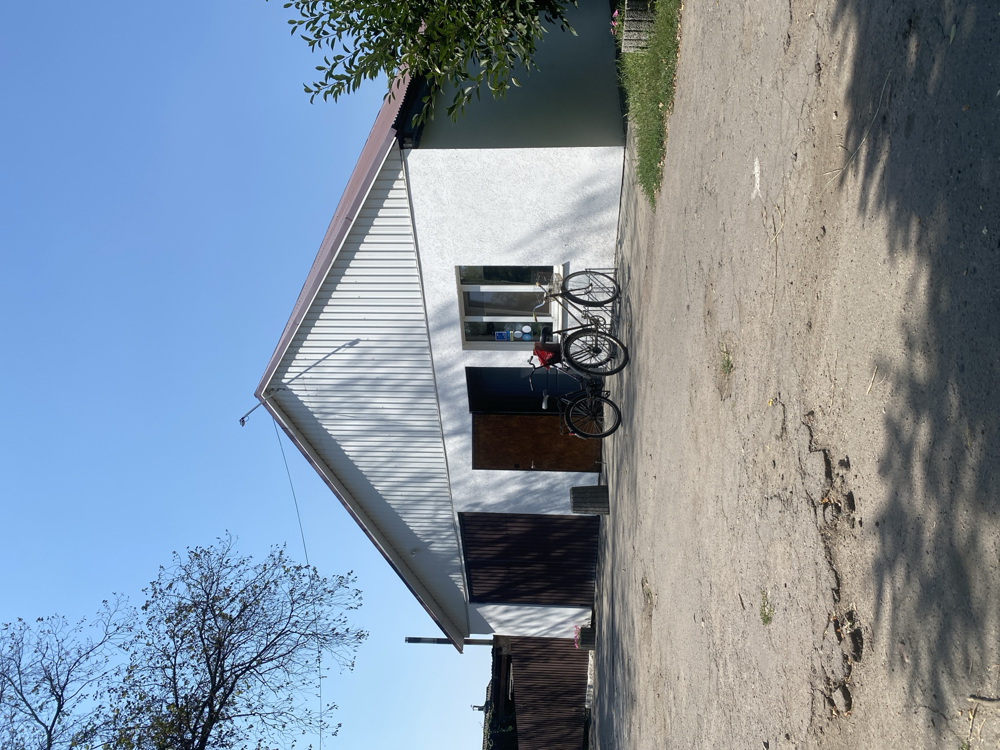 A sunlit, outdoor scene depicts a white, industrial-style building with a brown metal roof. The building has a large, dark brown double door on the left and a smaller, open wooden door to the right of it. To the right of the smaller door, there is a window with two panes. Two bicycles, one black and one black with a basket, are parked side-by-side in front of the building. The ground in front of the building is paved with a rough, gray asphalt surface that shows signs of wear and has several cracks and potholes. A dark brown wooden fence runs along the left side of the scene, partially obscured by a leafless tree branch in the upper left corner. In the upper right corner, the foliage of a green tree is visible. The sky is clear and blue. The location is identified as Ustyvytsia, Ukraine.