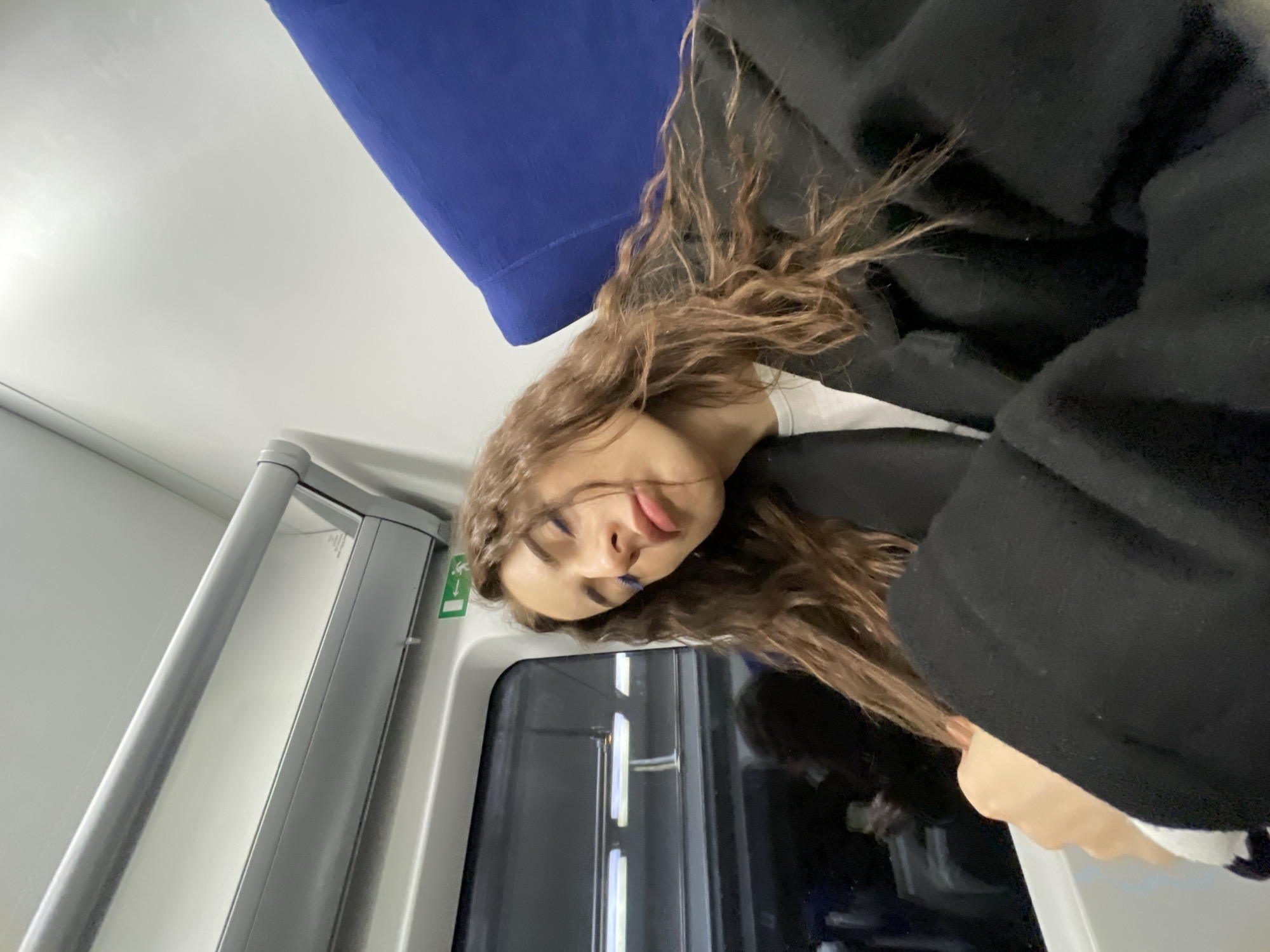 A young woman with long, wavy brown hair, wearing a black jacket and a white top, is posing for a selfie. Her face is close to the camera, and she is sticking her tongue out playfully. She has blue makeup on her eyelids. The background suggests she is inside a train car. A blue seat cushion is visible behind her head. A silver vertical bar and a train car door are also visible. Based on the style and color scheme of the train car interior, specifically the blue seat and gray interior, the location is likely a train in Moscow, Russia.