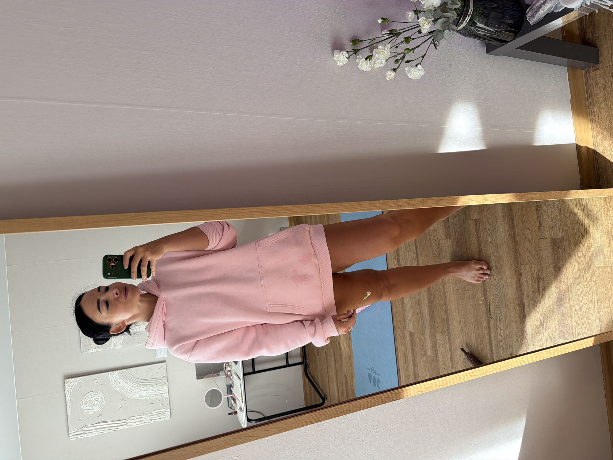The image is a mirror selfie of a woman wearing a light pink hooded sweatshirt. She is holding a green smartphone and appears to be taking the photo.
The reflection shows part of a room with light-colored walls. On the wall to the left is a piece of textured artwork. There is a desk with a vanity mirror and various items. On the floor, there is a blue yoga mat and a small brown object.
A portion of the room's ceiling is visible, adorned with white flowers in a vase. The floor appears to be made of wood.
The location cannot be determined with the information available.