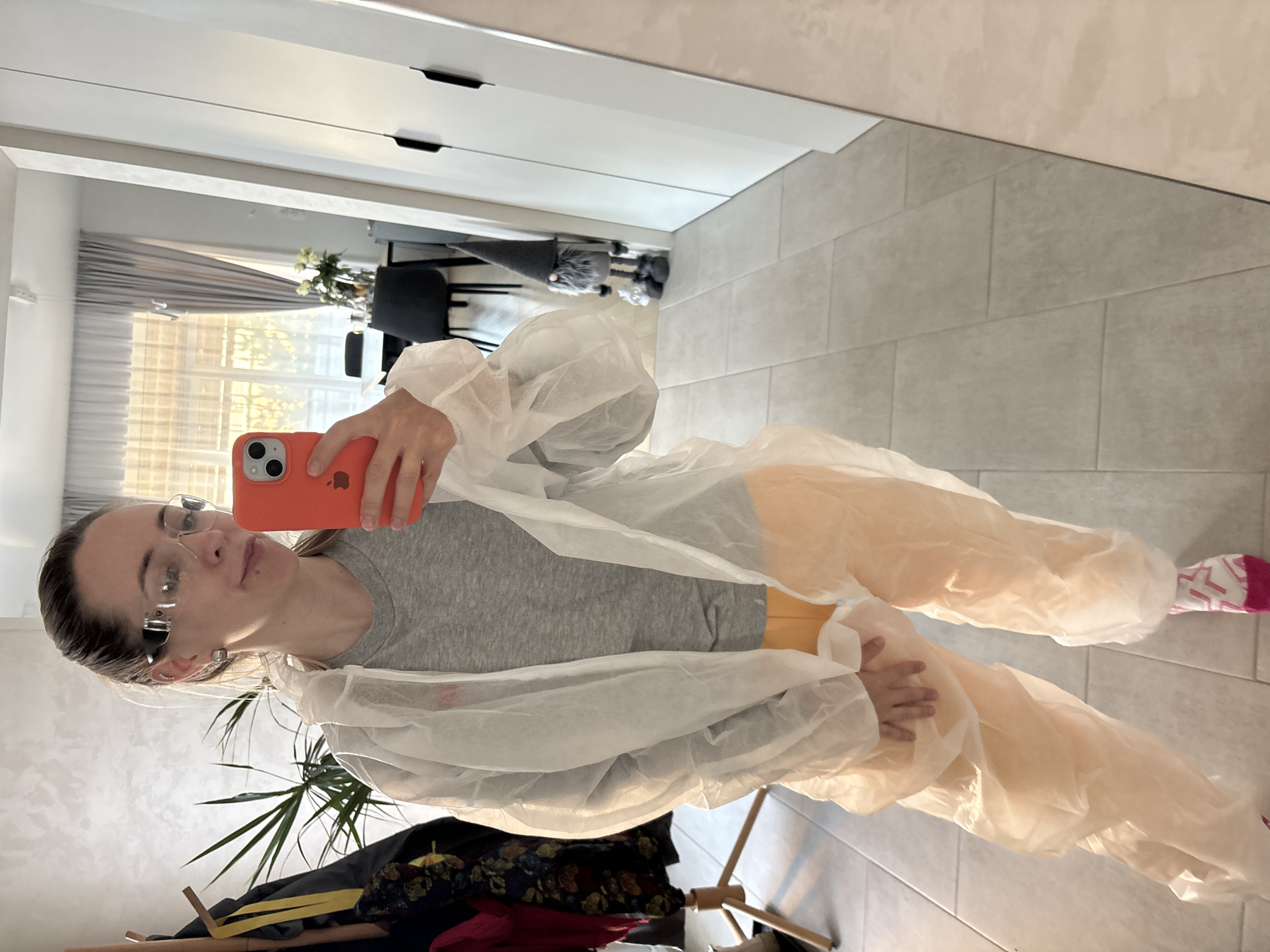 A person with glasses and a grey shirt is taking a selfie in front of a mirror. The person is wearing a white disposable coverall. The person is holding an orange phone. In the background is a plant, a coat rack with clothing, and a window with curtains. The scene is indoors, likely inside a residence. The location is Chaikovsky, Russia.