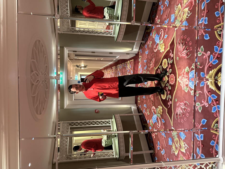 A male individual is captured in a full-length mirror selfie within a luxurious interior space. The subject, appearing to be in his 30s-40s with short, dark hair, wears a red long-sleeved Henley shirt, dark trousers, and black dress shoes. He holds a black smartphone in his right hand, angled for photography, and a small yellow-and-black object, possibly a key card, in his left hand. He stands on a distinctive red carpet adorned with intricate floral and botanical patterns in blue, green, yellow, and pink hues. The mirrored wall features ornate white frames with lattice designs, separated by vertical strips with crystal-like embellishments. To the subject's left and right are elegant console tables; one to the left holds a textured green decorative object. Behind the subject, a well-lit corridor extends, lined with light-colored doors, wall sconces, and a visible emergency exit sign. The white ceiling displays an elaborate circular ceiling medallion directly above the subject's reflection. A red fire alarm call point is visible on the wall to the left. The highly distinctive carpet and ceiling designs identify the location as a corridor within Claridge's Hotel in Mayfair, London, United Kingdom.
