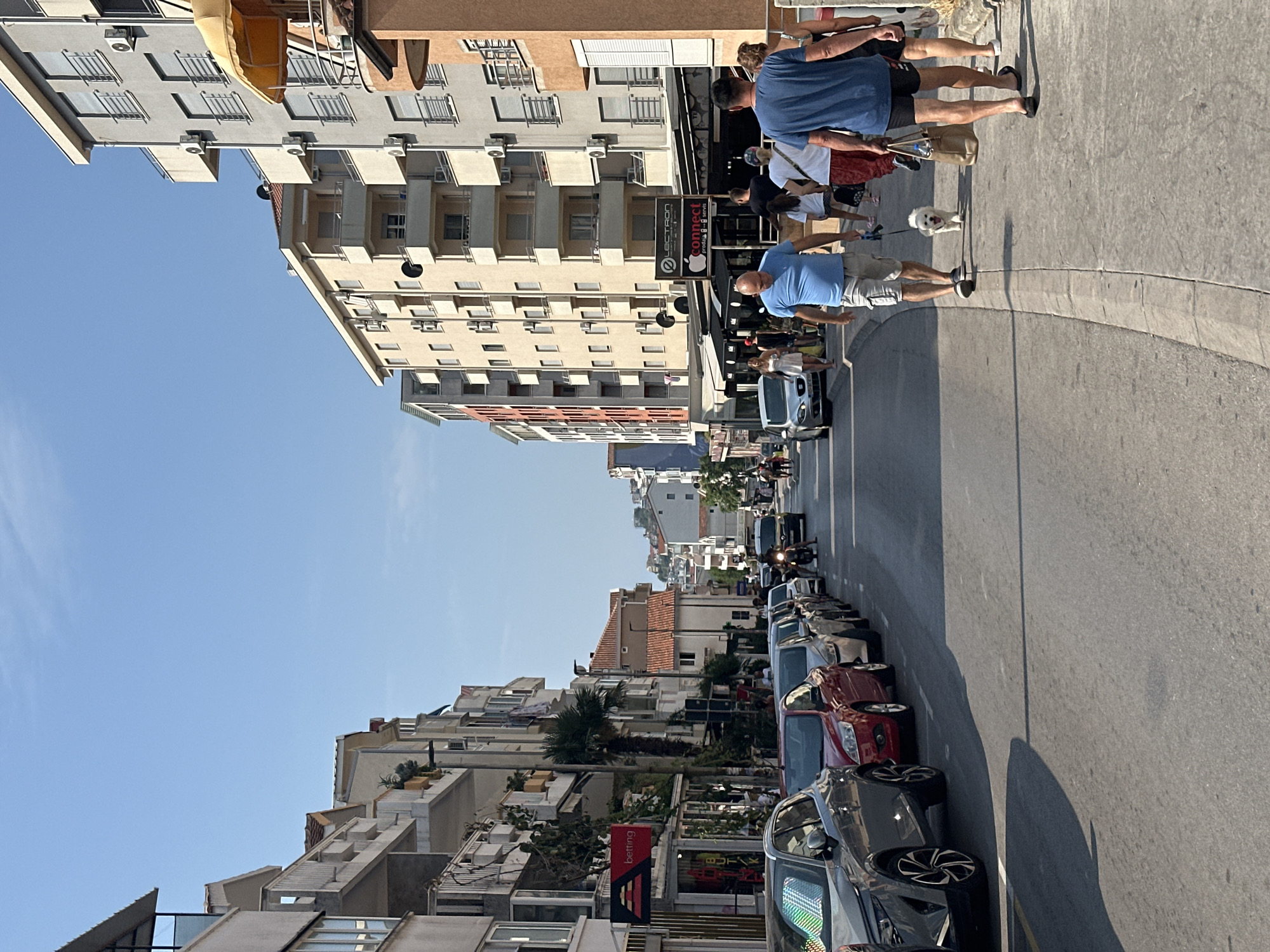 The image depicts a street scene in Budva, Montenegro, on a clear, sunny day. The street is lined with multi-story buildings, many of which feature balconies and air conditioning units. The architecture appears to be a mix of modern and older styles. The street itself is paved and has a pedestrian sidewalk on the right side.
Several cars are parked along the left side of the street, with one car in motion on the right side, possibly a motorcycle. People are walking on the sidewalk and in the street. On the right sidewalk, a man in a blue t-shirt and shorts is walking a small white dog on a leash. Behind him, another man in a blue t-shirt and black shorts carries a brown bag. Other pedestrians are visible further down the street.
The environment is urban, with shops and businesses visible on the ground floors of the buildings. The sky is clear and blue, indicating good weather. The overall impression is of a lively, populated urban area, likely a commercial or residential street in a coastal town.