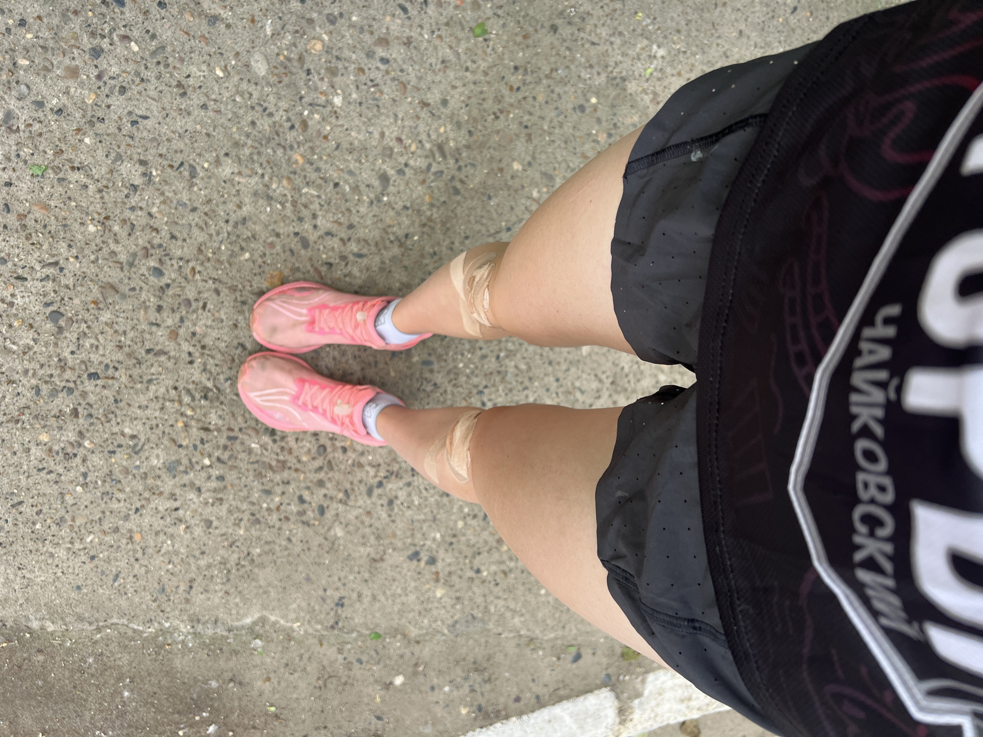 The image shows a person's legs and feet from a top-down perspective, standing on a concrete surface. The person is wearing bright pink athletic shoes with white laces and socks, black athletic shorts, and a black garment with a silver and purple design. There are two pieces of athletic tape applied to the front of each knee. The concrete surface appears to be a sidewalk or pavement, with small stones and debris visible. Part of a white curb is visible on the left side of the frame. The environment suggests an outdoor urban setting, possibly in Chaikovsky, Russia, as indicated by the lettering on the garment which appears to be in Cyrillic script, potentially spelling "Чайковский" (Chaikovsky).