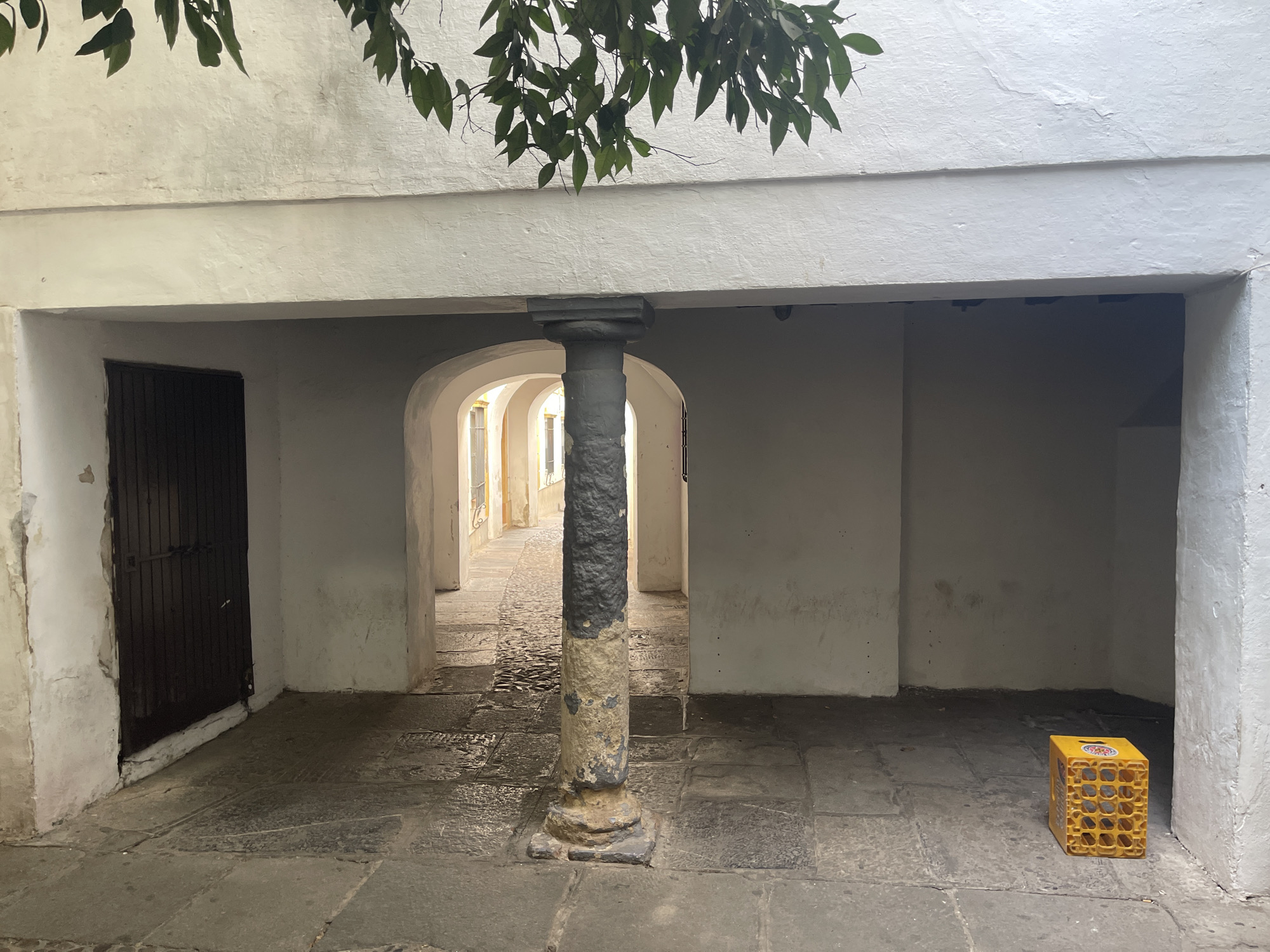 The image depicts a narrow, arched passageway with cobblestone flooring. A thick, cylindrical column, partially weathered and painted in dark grey and beige, stands centrally in the foreground. To the left, a dark wooden door with metal bars is visible. The passageway recedes into the distance, showing more arches and illuminated by natural light, suggesting an outdoor area beyond. Hanging from the top of the frame are dark green leaves, possibly from a tree. In the lower right corner, a yellow plastic crate, likely for beverages, sits on the cobblestones. The architecture and paving stones are typical of the historic Judería (Jewish Quarter) of Córdoba, Spain. The scene appears to be an outdoor courtyard or alleyway, illuminated by ambient light. No people are present.
