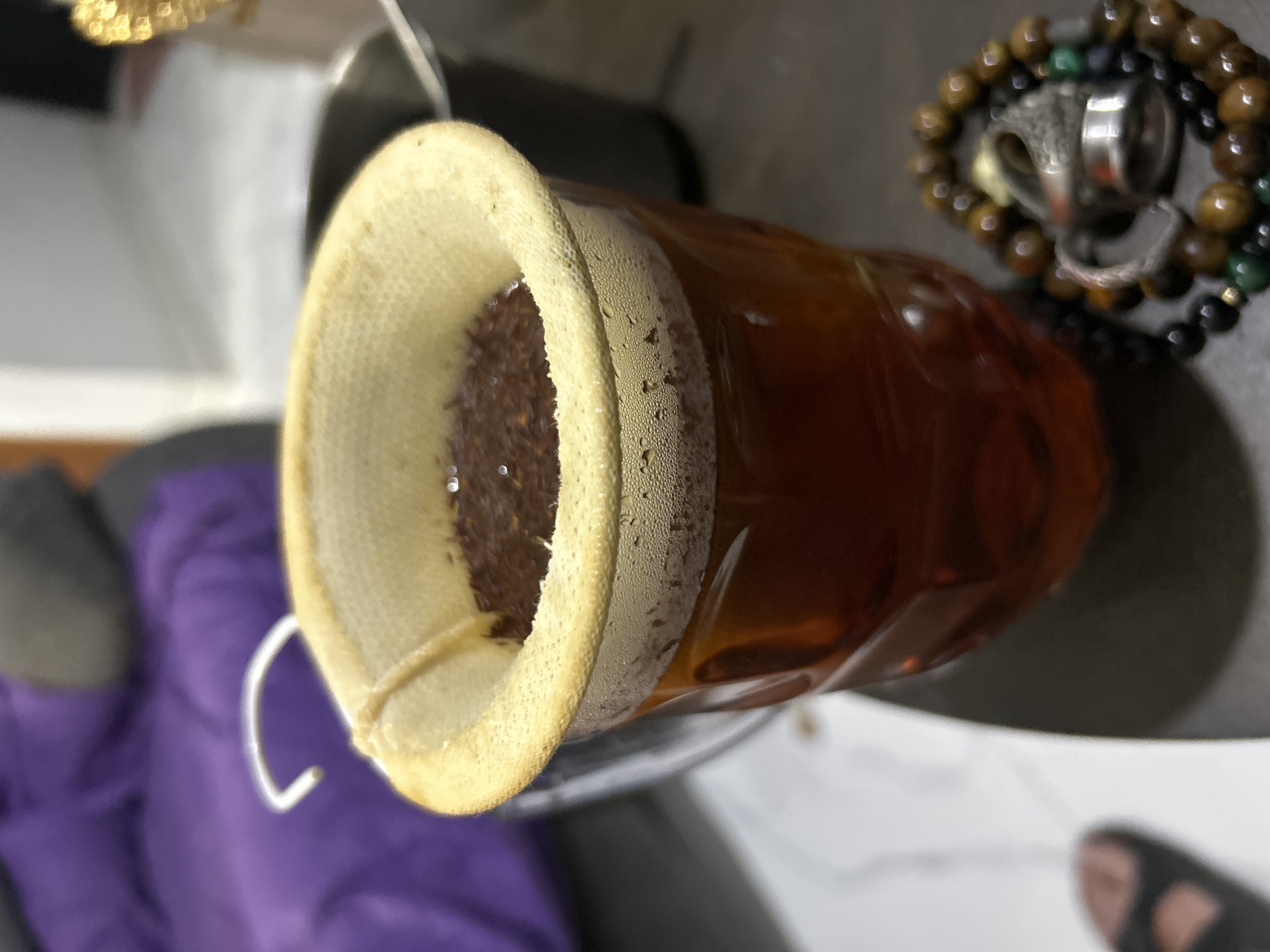 The image shows a close-up, top-down view of a beverage being strained through a fabric filter into a transparent container. The beverage is a deep amber color, likely tea or coffee, with small particles of the grounds visible at the top of the filter. The filter itself is made of a light-colored fabric with a reinforced rim. Condensation is visible on the outside of the container, suggesting it may be cold.
In the background, to the right, a stack of beaded bracelets is visible, one of which appears to be made of tiger's eye stones. There is also a ring with intricate detailing. To the left and bottom of the image, a blurred section of purple fabric is present. The overall setting appears to be indoors, with a focus on the preparation or serving of a beverage. There is no visible text in the image. The lighting suggests it could be daytime, but the exact time or weather conditions are not discernible. The image captures a simple, everyday activity.