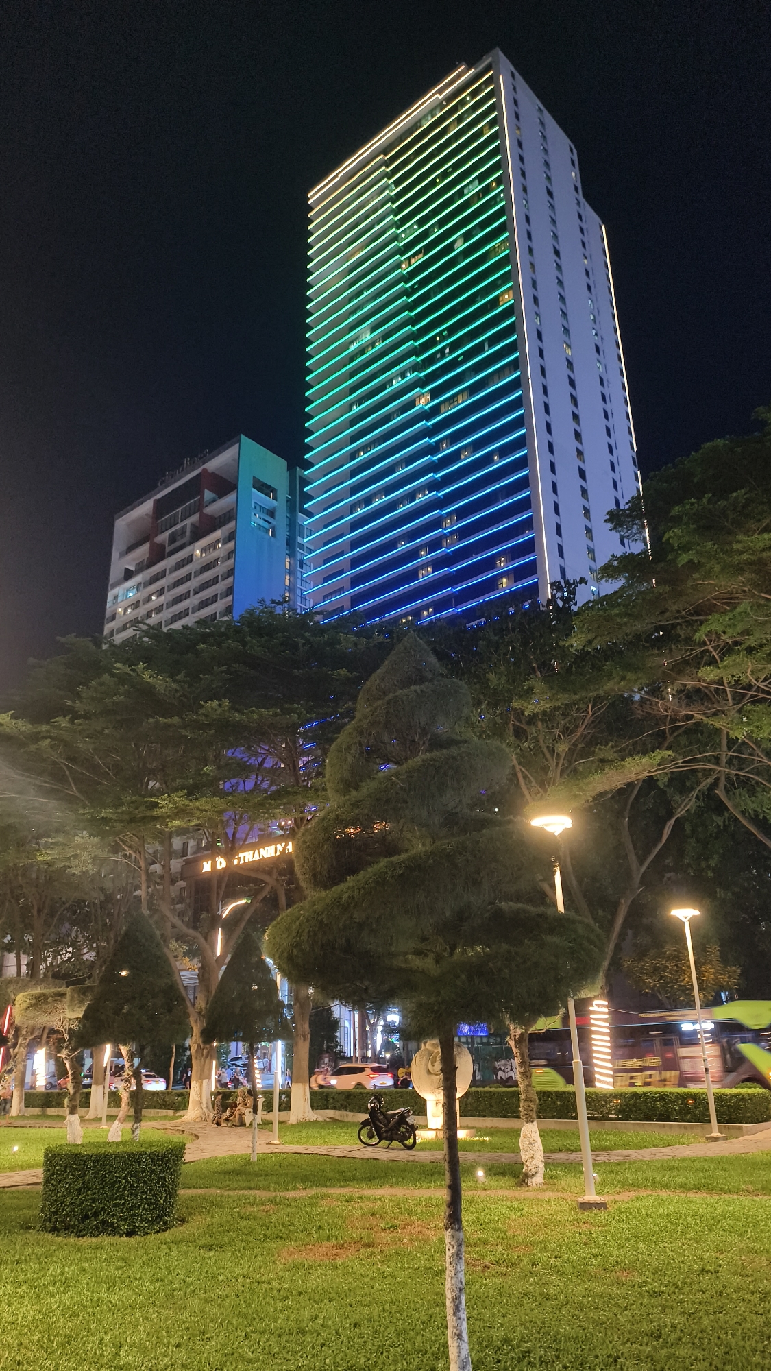 This image captures a nighttime scene in Nha Trang Ward, Vietnam, focusing on a modern, illuminated high-rise building. The building is predominantly white with a striking facade adorned with horizontal lines of lights that transition from yellow at the top to green and then to blue at the bottom. Adjacent to this is another building with a red and white exterior, also featuring blue lighting. In the foreground, a well-maintained park area features meticulously shaped trees, including a prominent spiral-shaped topiary. The ground is covered in green grass, with paved pathways winding through the park. Several streetlights with a warm glow are visible. In the background, traffic is present, with a bus and several cars visible, as well as a motorcycle parked on the side. A few individuals are seated in the park area. The overall atmosphere is urban and vibrant, highlighted by the colorful illumination of the buildings against the dark night sky.