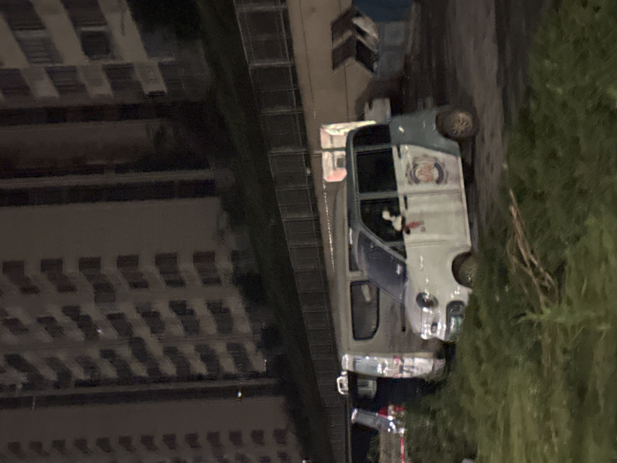 The image depicts an outdoor scene at night, likely in the Chongqing High-tech Zone, China. A multi-story building with numerous windows is visible in the background, suggesting an urban or developed area. In the foreground, several vehicles are parked. A small, light-colored vehicle with a blue stripe and a circular emblem on its side is centrally positioned. Adjacent to it, a larger white van is parked. To the left, a dark-colored car is visible. The ground appears to be covered with grass and some debris. In the mid-ground, a metal fence runs horizontally. Further to the right, two large blue trash receptacles are open, with some white objects visible inside. The lighting conditions suggest it is nighttime, with artificial light sources illuminating the scene, casting some reflections on the vehicles.