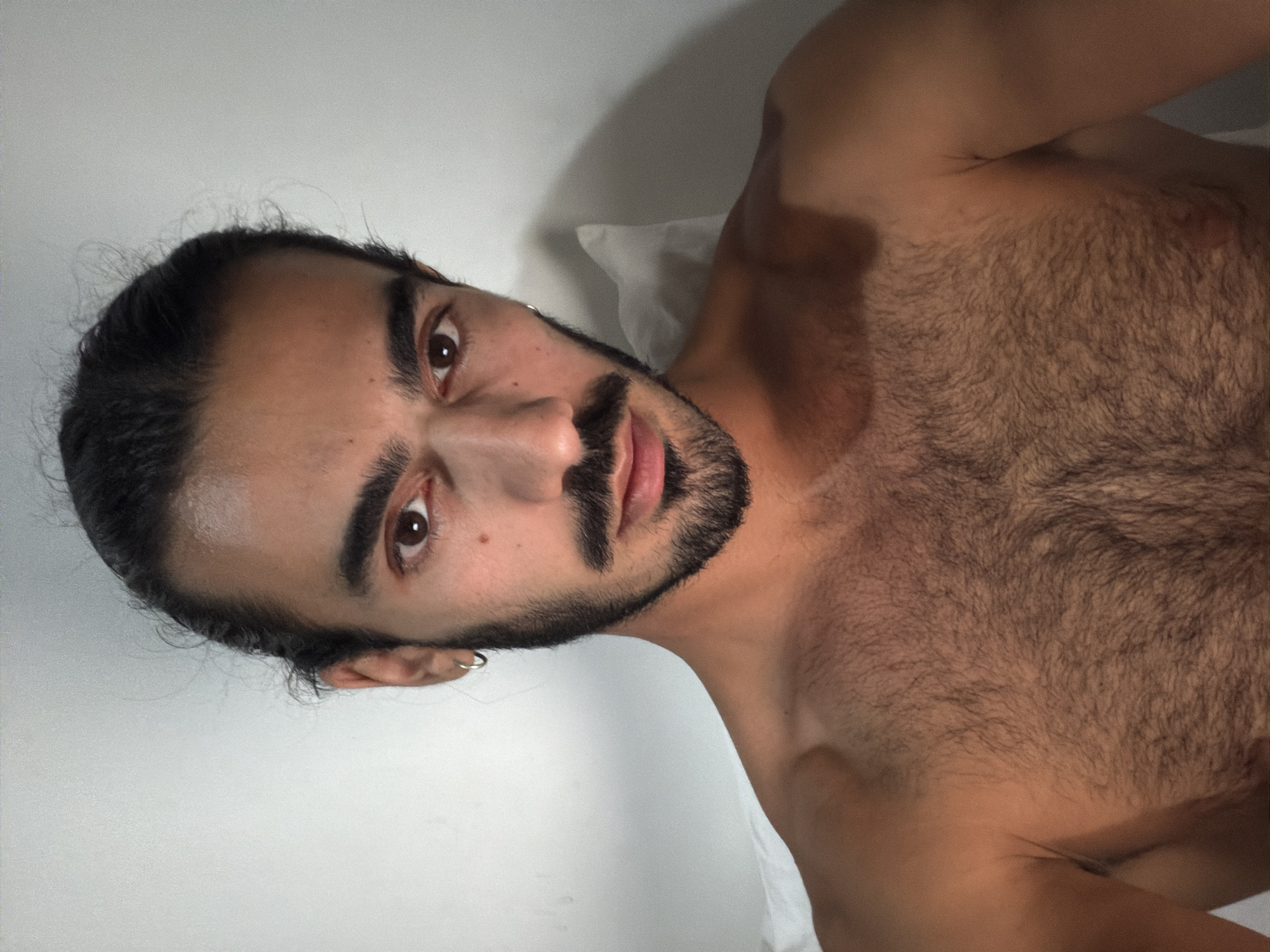 The image is presented on its side, depicting a man reclining and looking directly at the camera. He has dark hair, pulled back, and a prominent dark beard and mustache. His dark brown eyes engage the viewer with a direct and neutral expression. A small silver hoop earring is visible in his left ear. The man's upper torso and chest are bare, displaying dense dark body hair. He appears to be lying on white bedding, likely a pillow, which supports his head and shoulder. The background is a plain, light-colored, possibly white, wall or surface. The lighting is soft and diffused, without harsh shadows, suggesting an indoor setting. No text is visible in the image. The photo's context indicates it was taken in North Kuta, Indonesia.
