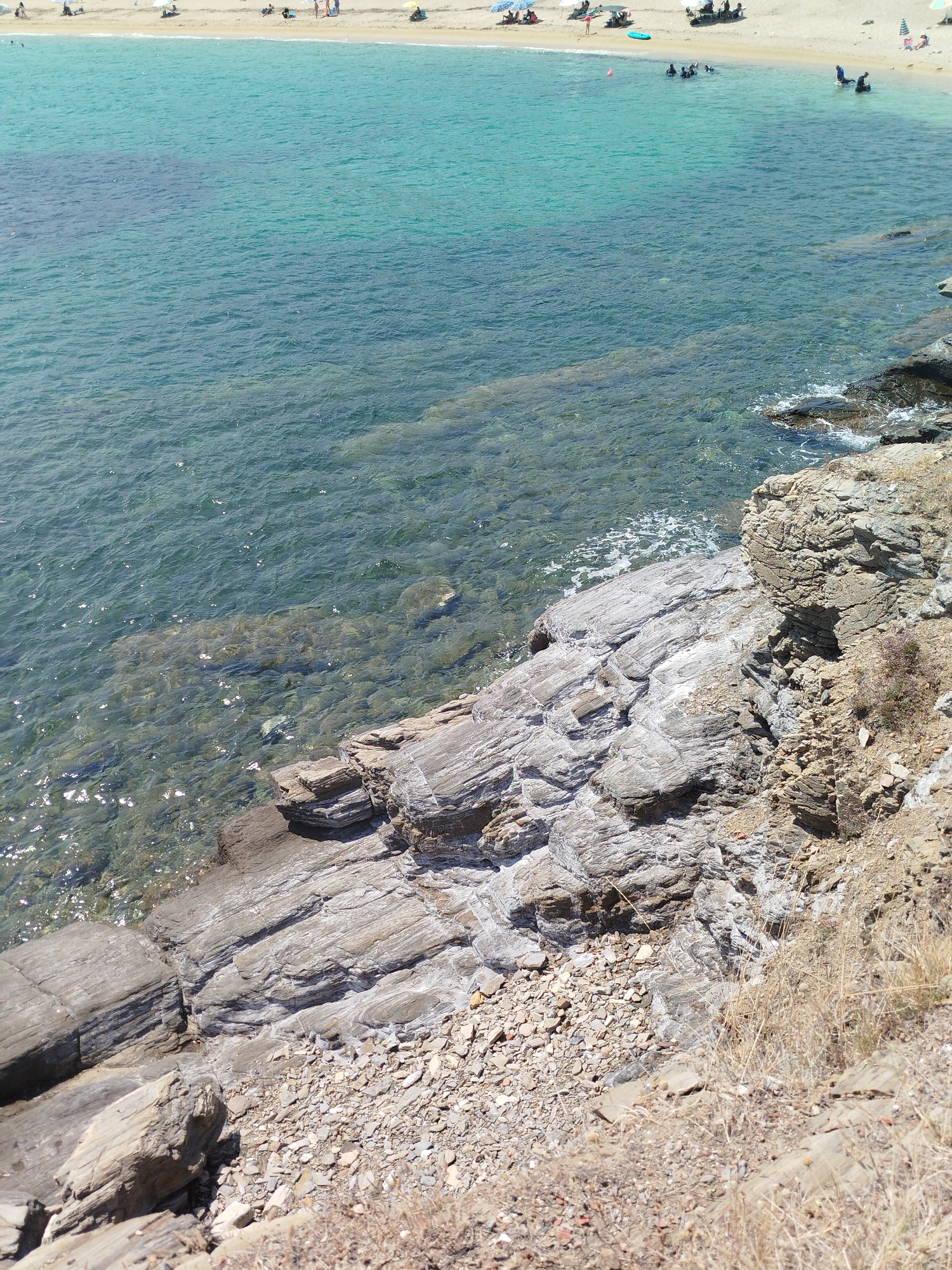This is an eye-level, medium shot of a rocky shoreline with clear, turquoise water in the foreground and a sandy beach with people in the background. The rocks in the foreground are layered and weathered, with some dry vegetation and small stones scattered on them. The water is shallow and transparent, allowing the seabed to be visible in places. The beach in the distance is populated by several individuals, some sitting under umbrellas, others walking or swimming. The scene depicts a typical summer day at a coastal location. This image is consistent with views found along the coastline of Sithonia Municipal Unit, Greece, known for its clear waters and sandy beaches.