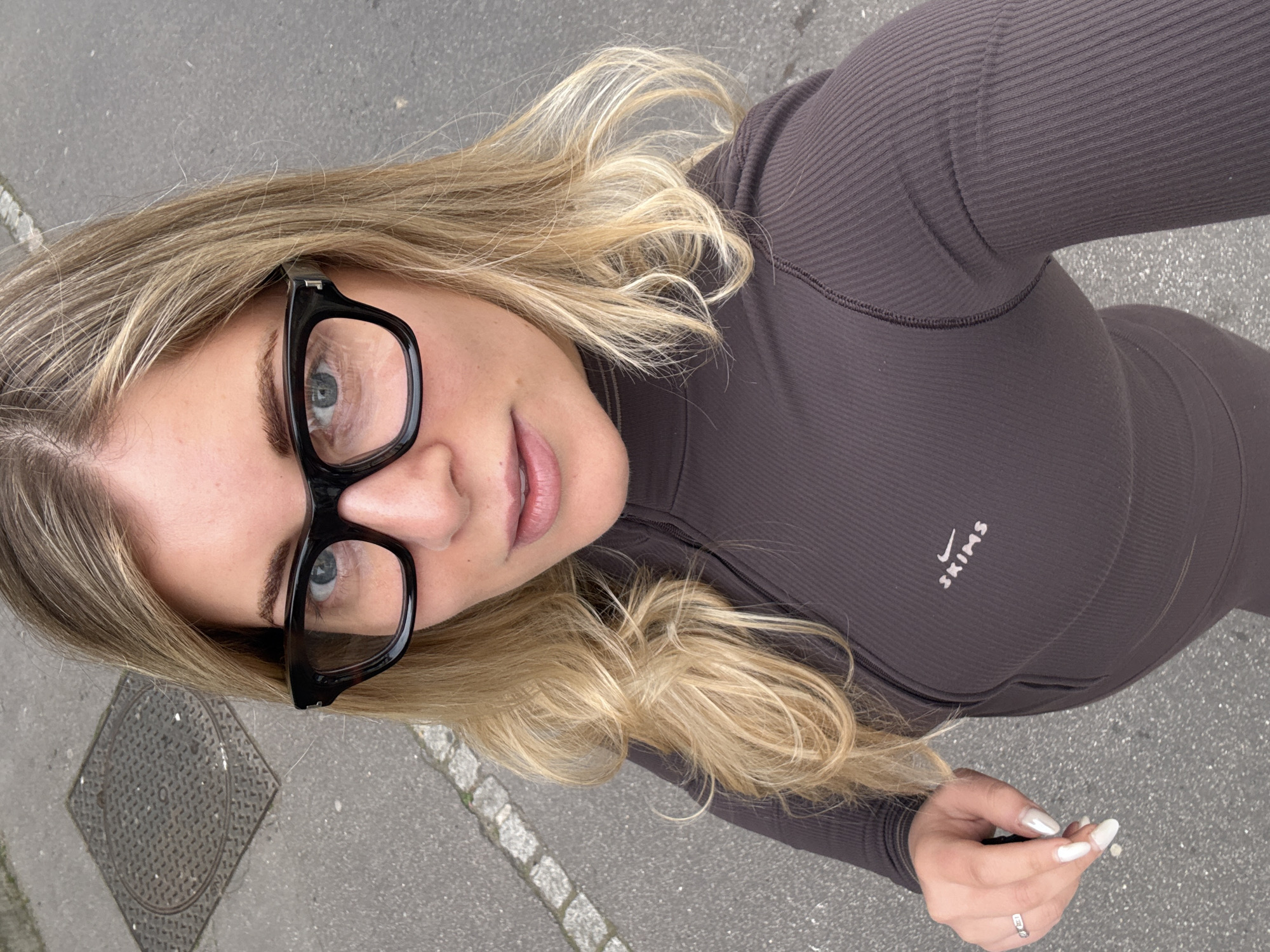 An overhead, slightly rotated photograph captures a young woman outdoors in what appears to be a city setting, likely Paris, France. She is looking upwards directly at the camera. Her blonde hair, styled with loose waves, cascades around her face and over her shoulders. Her eyes are a striking blue, framed by thick black-rimmed glasses that sit prominently on her face. Her eyebrows are well-defined, and her lips have a natural, soft pink hue.
She is wearing a dark brown or grey ribbed long-sleeved top, which appears to be part of an athletic or loungewear set, possibly extending to matching pants, visible in the lower portion of the image. The brand name "SKIMS" is vertically printed in white on the chest area of her top, accompanied by a small white logo resembling a Nike swoosh. On her left hand, which is visible in the lower right, she wears a silver ring on her ring finger, and her fingernails are painted in a long, pointed style with a white or iridescent finish.
The background consists of grey asphalt or pavement, suggesting a sidewalk or street. A square metal utility plate or manhole cover with a circular pattern is visible on the ground beside her head. The bright, even lighting indicates it is daytime with clear weather. The overall impression is a casual, outdoor portrait taken from a high angle.