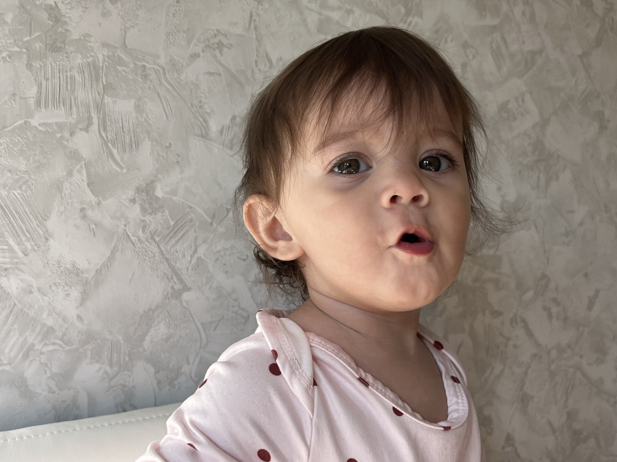 A toddler with brown hair and brown eyes, wearing a pink shirt with brown polka dots, is looking at the camera with a surprised expression. The toddler's mouth is open. The background is a gray textured wall. The city and country are not identifiable from the visual information.
