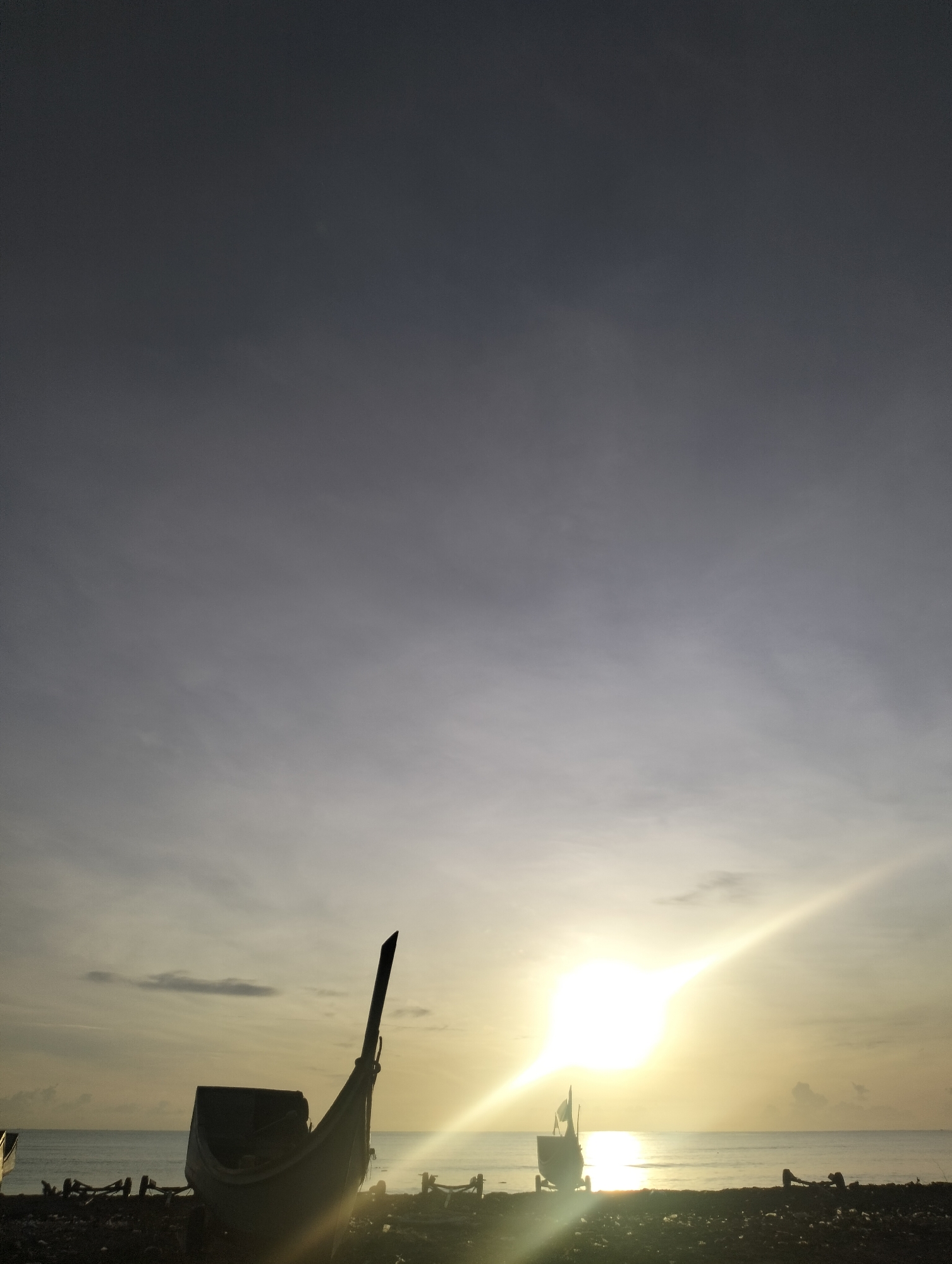 The image depicts a tranquil beach scene in Calok, Indonesia, during what appears to be either sunrise or sunset. The sky is a gradient of dark gray at the top, transitioning to lighter shades of gray and yellow towards the horizon, with soft clouds scattered across the expanse. The sun is a bright, glowing orb, positioned low on the horizon, casting a strong lens flare that stretches across the sky.
In the foreground, silhouetted against the bright sky, are several traditional Indonesian fishing boats. One prominent boat, positioned slightly to the left of center, is tilted on its side, its hull and pointed prow clearly visible. Its dark shape contrasts sharply with the luminous sky behind it. Other, less distinct boat shapes and what appear to be wooden stands or frames are scattered along the dark, pebbly shore.
The ocean stretches out to the horizon, reflecting the soft light of the sun, creating a shimmering effect. The water appears calm, with a gentle line where it meets the shore. The overall atmosphere is serene and peaceful, suggesting the quiet end or beginning of a day for local fishermen. No people are visible in the image, emphasizing the solitude of the location. The environment is characterized by its natural elements: sky, sea, and land, with man-made objects (the boats) harmoniously integrated into the landscape.