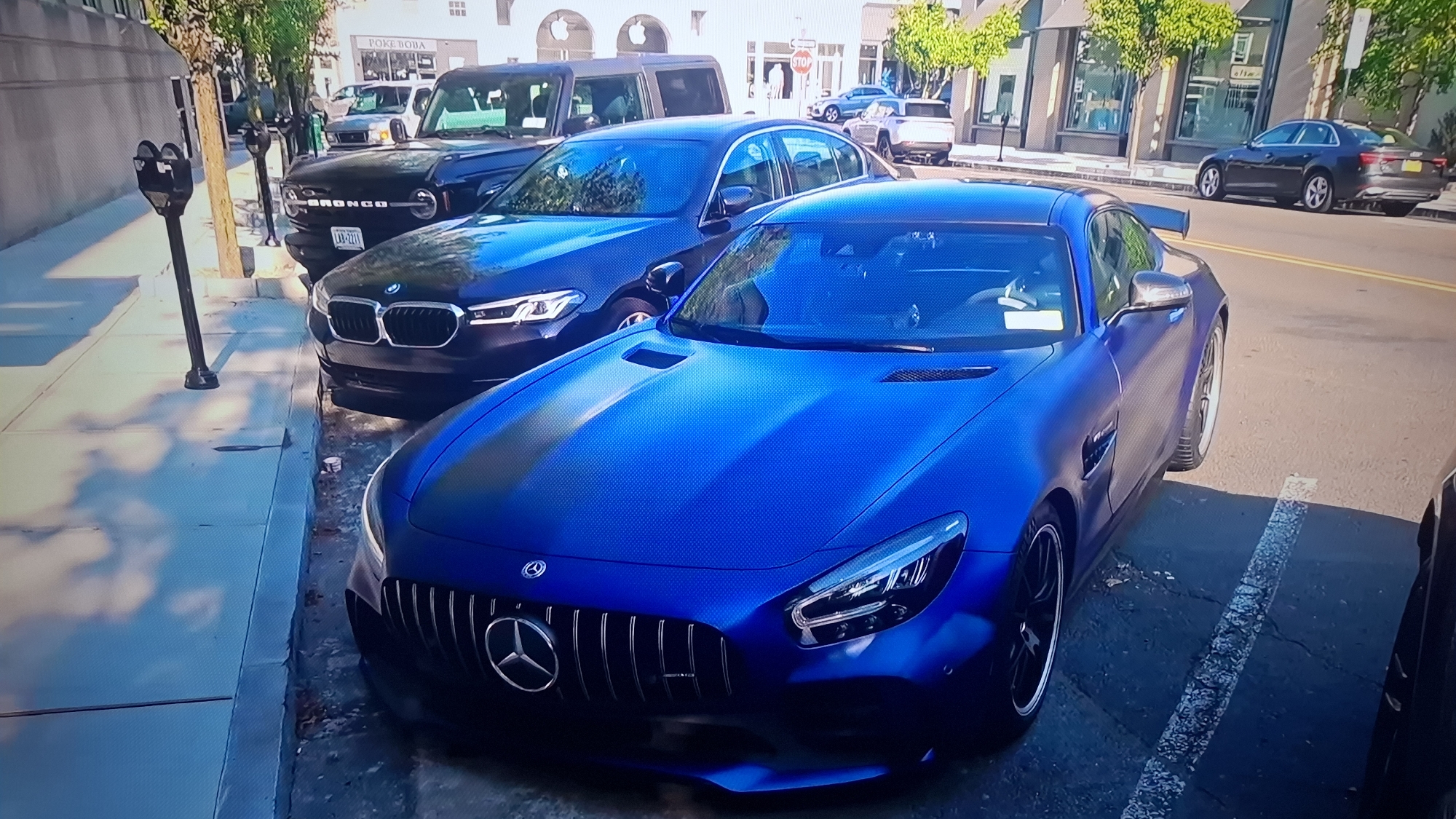 The image shows a street in an urban area, with several parked vehicles. The foreground features a blue Mercedes-AMG GT with a large front grill and distinctive headlights. Behind it is a dark blue BMW sedan, partially obscured. Further back is a black Ford Bronco SUV. In the background, other vehicles are parked along the street, including an Audi sedan. The street appears to be well-maintained, with a sidewalk on the left side of the frame. A parking meter stands on the sidewalk next to the Mercedes. Buildings are visible in the background, with recognizable storefronts such as "POKE BOBA" and a store with an Apple logo. The lighting suggests it is daytime. The location is Unknown, Unknown.