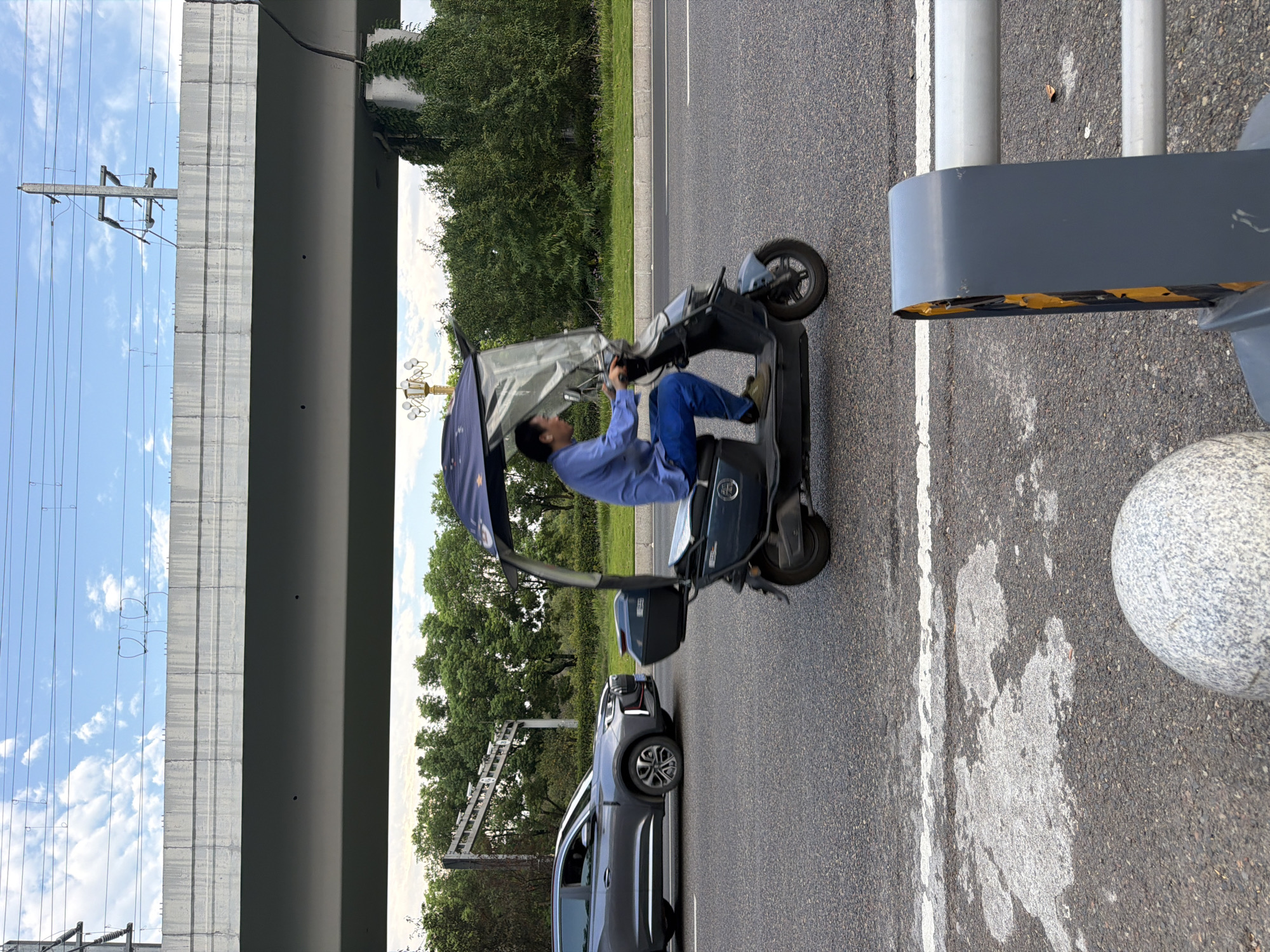 A man wearing a blue shirt and blue pants is riding a dark gray electric scooter with a clear canopy and a black storage box on the back. He is on a paved road with white lane markings. To the left of the scooter, a gray SUV is driving in the same direction. In the background, there are green trees and a concrete overpass with electrical wires and poles. The sky is cloudy with patches of blue. The scene appears to be in Jiangjin, China.