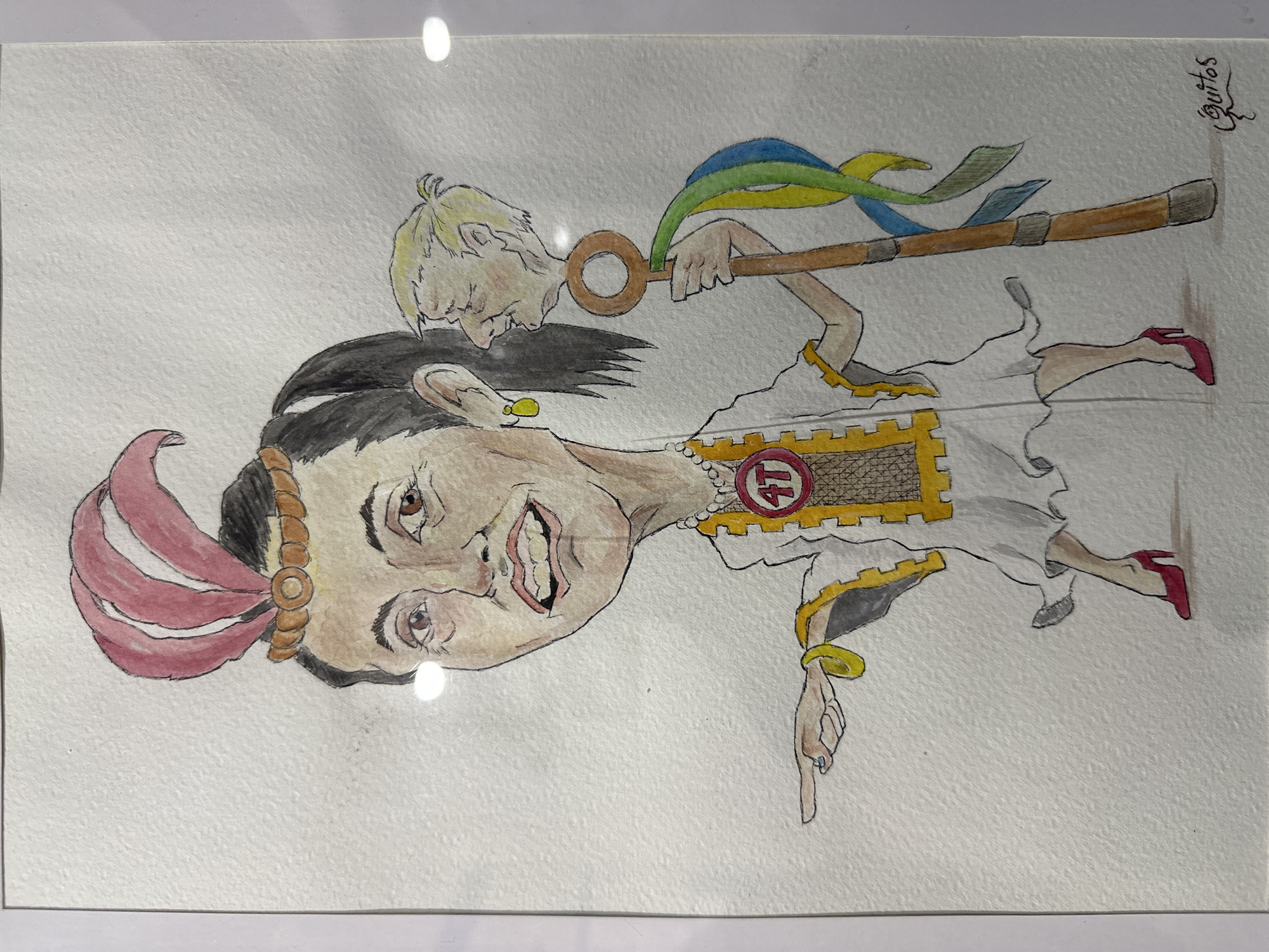 A framed watercolor painting depicting a caricature of Claudia Sheinbaum, the current Head of Government of Mexico City, combined with elements of Aztec culture. Sheinbaum is shown wearing a headdress of red feathers and a white dress with a gold trim reminiscent of Aztec clothing. A golden number 4T is prominent on her chest, referencing the Fourth Transformation. An additional figure with blond hair holds a large key with ribbons of blue, green, and yellow attached. Sheinbaum points downwards. The artwork is signed “Quitos” in the upper right corner. The style of the painting is cartoonish and satirical. The location is implied to be Mexico City due to the subject matter.