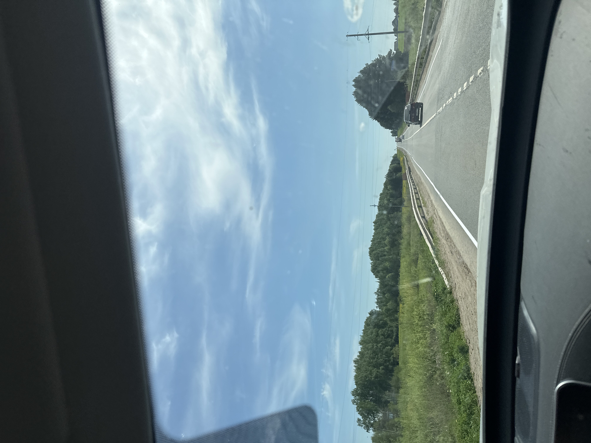 The image shows a view from inside a vehicle, looking out through the windshield. A highway stretches into the distance, with a black car driving ahead in the same lane. The sky is blue with scattered white clouds. Green trees and vegetation line the side of the road. The dashboard and frame of the car are visible on either side of the image. The location is possibly Nylga, Russia, judging by the geographical features of the road, trees and vegetation.