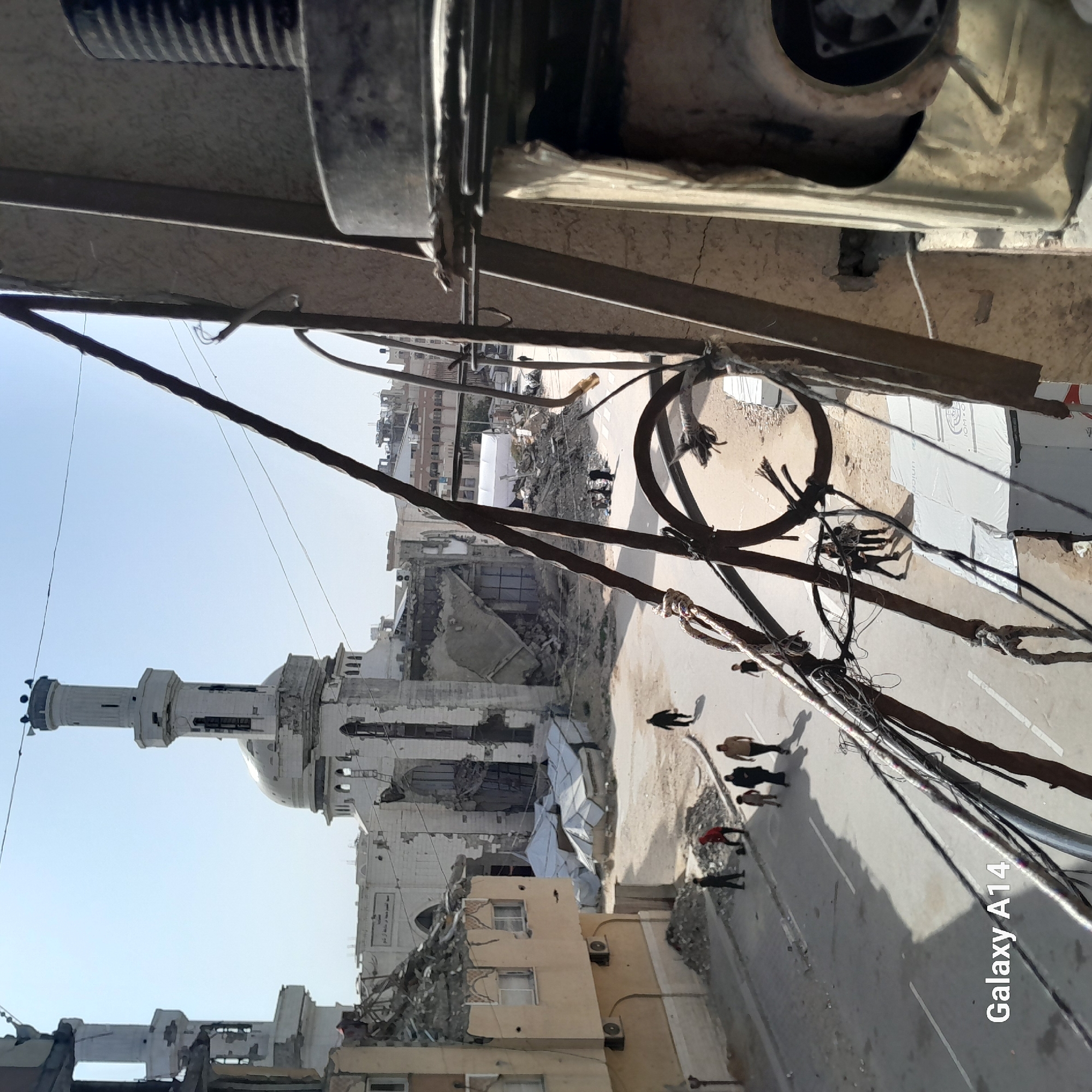 The image, taken from an elevated perspective, captures a scene of urban destruction under bright daylight. In the immediate foreground, numerous thick, dark cables and wires are strung across the frame, some bundled and knotted, suggesting utility infrastructure.
Below, a wide street with faint white lines is visible, lined by heavily damaged buildings and piles of rubble. A large, white mosque with a tall minaret and a prominent dome stands as a central feature, appearing partially damaged but largely intact against a clear, pale blue sky. Many surrounding structures are severely compromised, with exposed interiors and collapsed sections, indicating widespread destruction.
Several individuals are dispersed on the street below, some walking alone and others in small groups, casting distinct shadows that confirm a sunny day. One group of people stands near a large, coiled cable on the street. Further down the street, a white, tent-like structure is present amidst the debris. On the wall of a building near the mosque, some indistinct Arabic text is visible. The bottom right corner of the image displays a "Galaxy A14" watermark in white text. The scene depicts daily life continuing amidst a severely damaged urban environment.