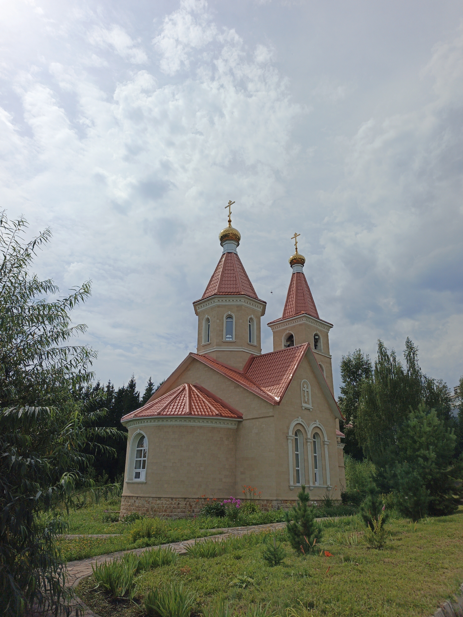 The image depicts a light-colored church with a red tiled roof, featuring two golden domes topped with crosses, and a bell tower also adorned with a cross. The church is situated in a park-like setting with lush green grass, various plants, flowers, and trees. A stone walkway meanders through the grass towards the church. The sky is partly cloudy with dramatic cloud formations. The architectural style and the presence of Orthodox crosses suggest the church is likely an Orthodox Christian church. Based on contextual information, this church is located in the city of Zlatoust, Russia.