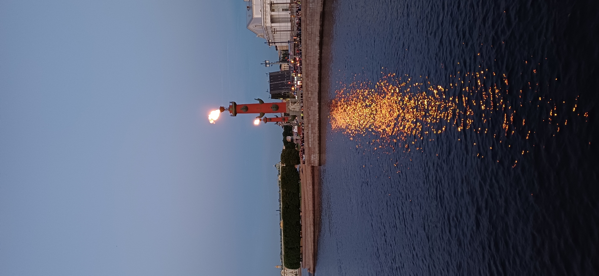 The image depicts a view of the Neva River in Saint Petersburg, Russia, at dusk. Prominently featured are two Rostral Columns on Vasilievsky Island, one of which is actively burning with a large flame. The other column also shows a smaller flame. In the background, several buildings, including a large, classical-style structure with columns and a white facade, are visible along the riverbank. A dense line of trees forms a hedge along the waterfront promenade. A crowd of people is gathered on the embankment, observing the columns. The dark water of the Neva reflects the vibrant orange flames, creating a shimmering pathway of light on the surface. The sky is a gradient of deep blue to a lighter blue near the horizon.