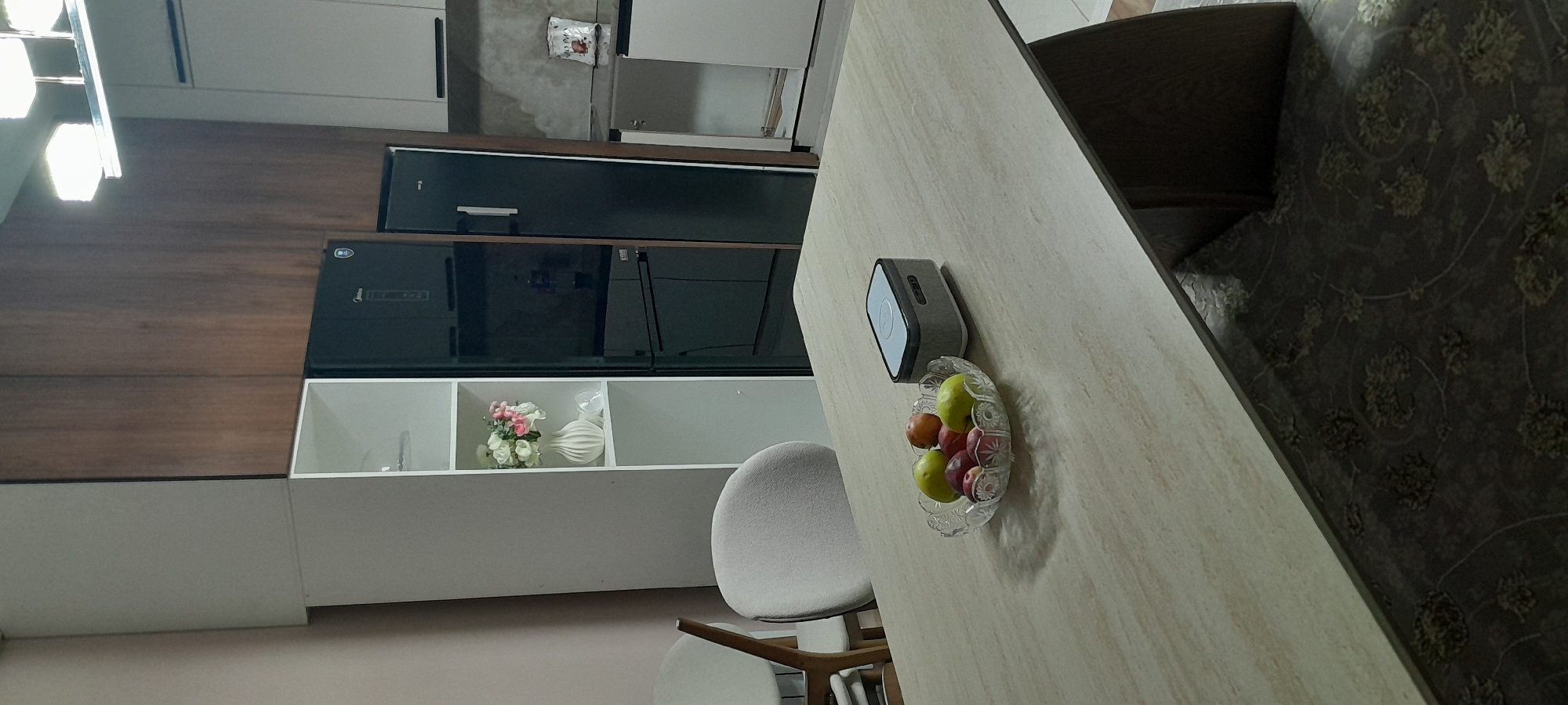 The image depicts an indoor scene, likely a dining or kitchen area. In the foreground, a light-colored, textured table holds a clear glass bowl filled with various fruits, including apples and possibly plums. To the right of the fruit bowl is a grey rectangular electronic device. In the background, a tall, white shelving unit is visible, containing a white vase with a bouquet of pink and white flowers. Adjacent to the shelving unit is a black refrigerator. Further to the right, kitchen cabinets are present. The overall environment suggests a modern, domestic setting. There is no indication of the city of Kyzyl-Asker, Kyrgyzstan.