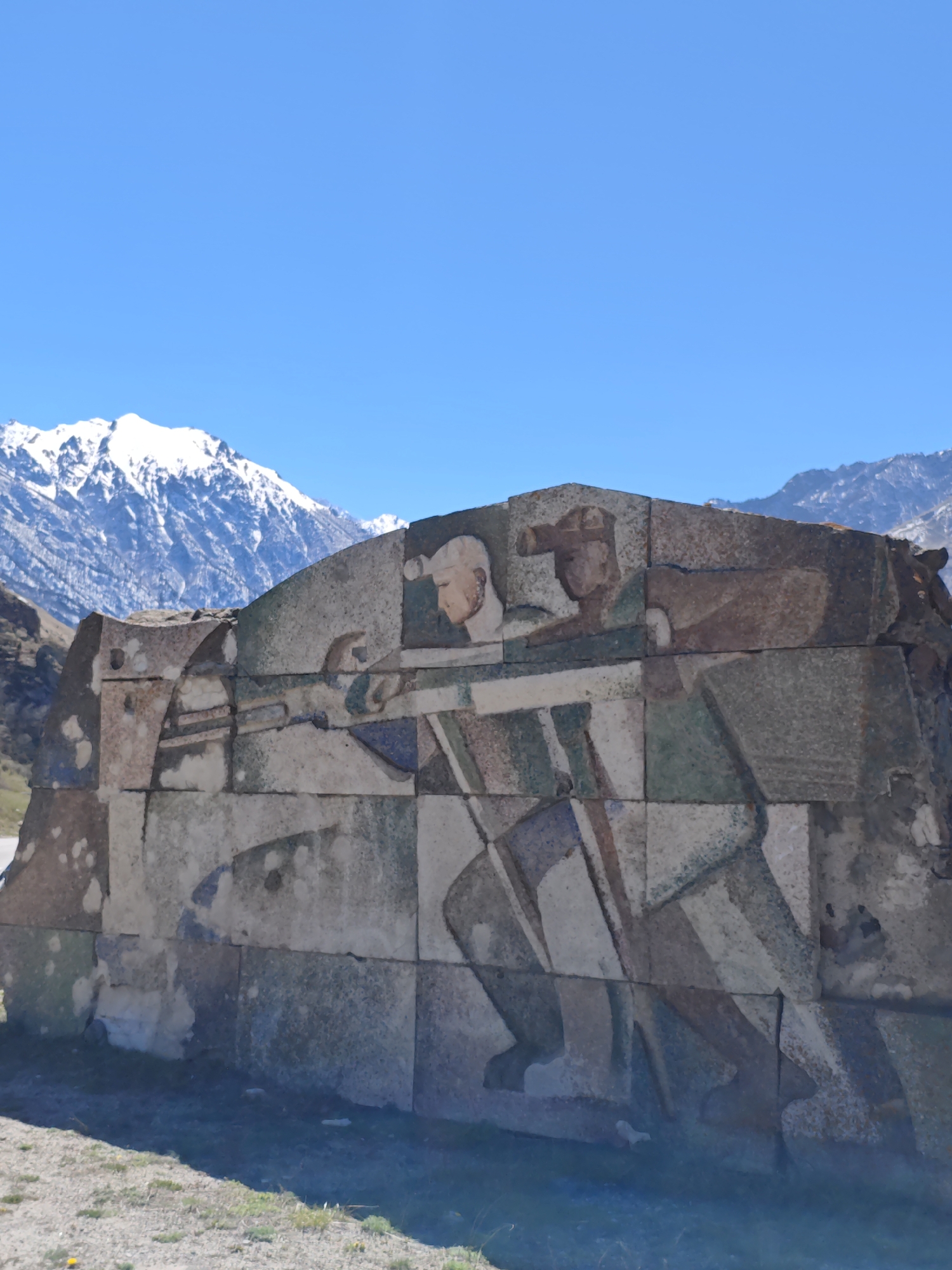 The image shows a large stone block monument featuring an image of two men with rifles, set against a backdrop of snow-capped mountains and a clear blue sky. The monument is located on a grassy area and appears to be in Tyrnyauz, Russia, based on the image's context. The figures are depicted in a stylized, somewhat abstract manner, possibly Soviet-era design. The monument itself is constructed of irregularly shaped stones and appears weathered, suggesting it is not new.