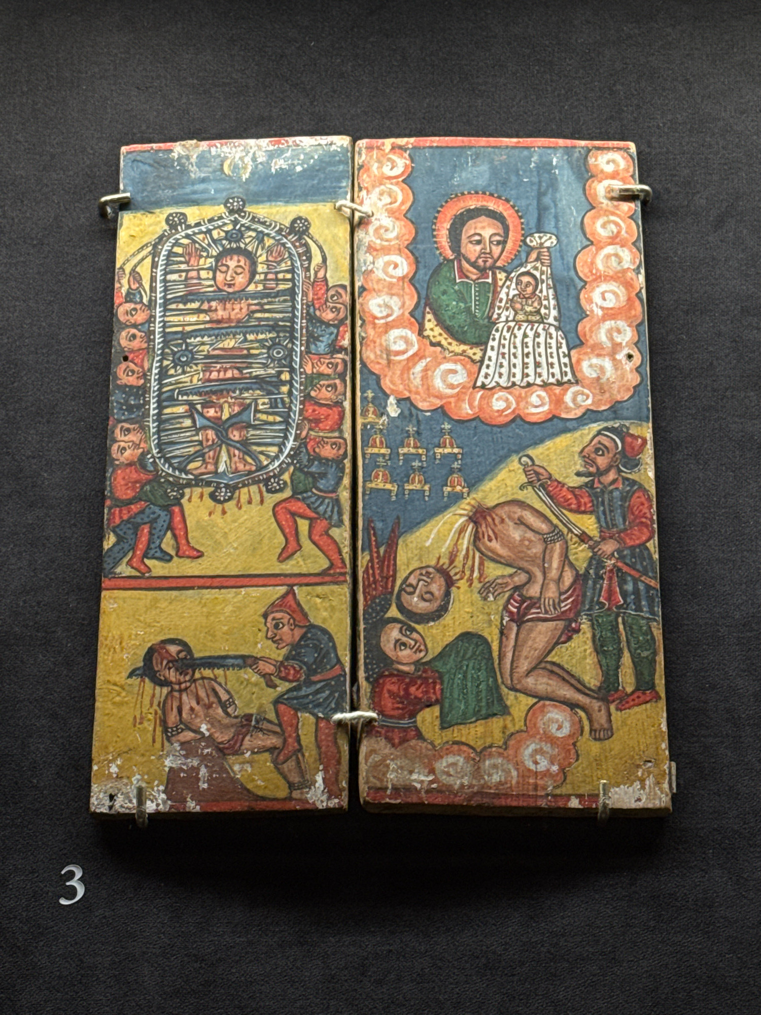 This is a photograph of a diptych, a two-paneled artwork, on a dark background. The diptych is made of wood and appears to be old, with some wear and tear.
The left panel depicts a scene with several figures. At the top, a person is seen in what appears to be a hanging or stretched position within an ornate frame, with onlookers below. The figures are stylized and painted in vibrant colors.
The right panel features a depiction of a man holding a child, possibly a religious icon. Below this, there is a scene of violence, with one figure decapitating another. Other figures are present in the scene, observing or participating in the events. The overall style of painting is folk art, with flat colors and a lack of perspective.
The object is placed on a dark, textured surface, possibly fabric or a dark wall. A white numeral "3" is visible in the lower left corner of the image, suggesting it might be part of a museum display or catalog. The location of this object in Moscow, Russia is not provided in the image or its accompanying information.