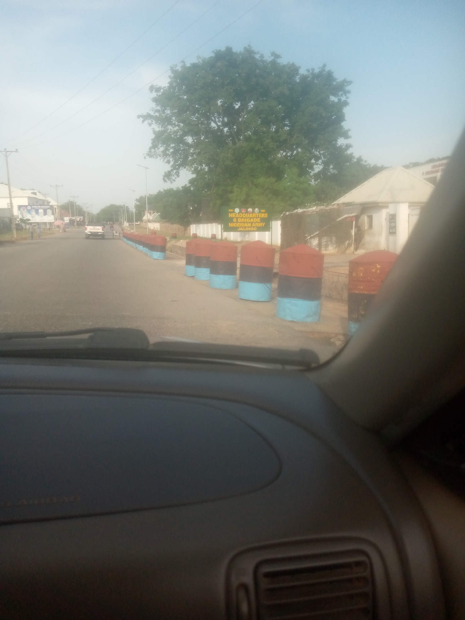 The image depicts a street scene in Jalingo, Nigeria, viewed from inside a car. The time of day appears to be late afternoon or early evening, with the sky a pale blue and the sun casting long shadows. The weather is clear.
The foreground is dominated by the interior of the car, showing the dashboard and part of the steering wheel. The road stretches ahead, lined with a series of cylindrical barriers painted in red, black, and light blue. These barriers are arranged along the side of the road, suggesting a security checkpoint or a traffic management measure.
In the middle ground, a white Toyota pickup truck is visible on the road, along with other vehicles and what appear to be pedestrians. To the right of the road, there is a building with a distinctive pitched roof, and some overgrown vegetation. Further down the road, there are more buildings and trees.
A prominent sign on the right side of the road reads "HEADQUARTERS 6 BRIGADE NIGERIAN ARMY JALINGO." This sign provides a clear indication of the location and the context of the scene. The overall atmosphere seems calm and everyday, with no obvious signs of distress or unusual activity. The scene is likely part of a routine day in Jalingo.