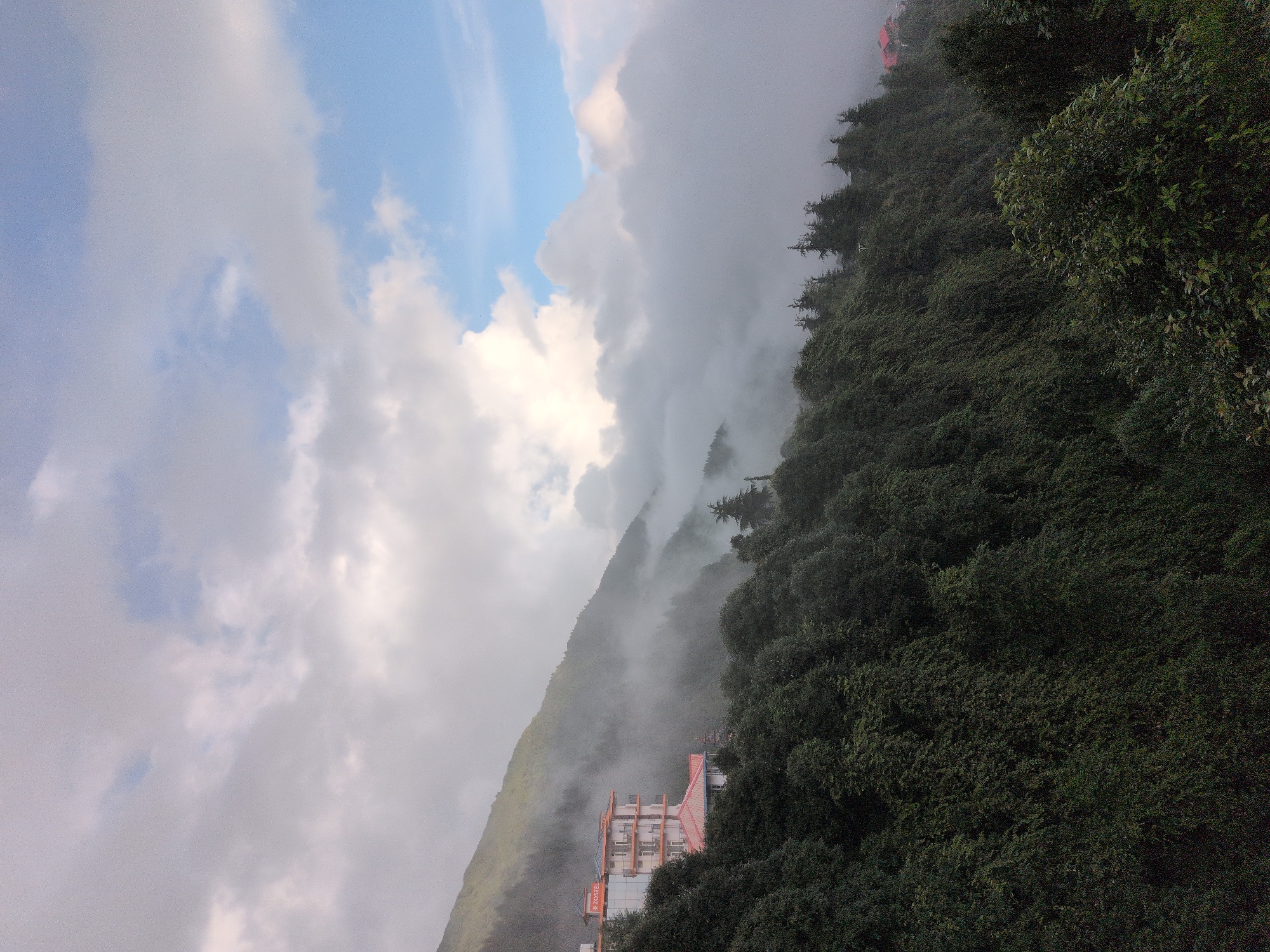 A hostel building with orange and white exteriors is nestled on a mountainside covered in lush green trees. The building has multiple floors with balconies and is partially obscured by fog that creeps up the mountain. The sky above is a mix of white clouds and patches of blue, suggesting either early morning or late afternoon. The landscape is mountainous and appears to be located in a remote, possibly hilly region in India, with the specific location being Haini, India. The image is taken from a slightly elevated perspective, looking up towards the building and the mountain.