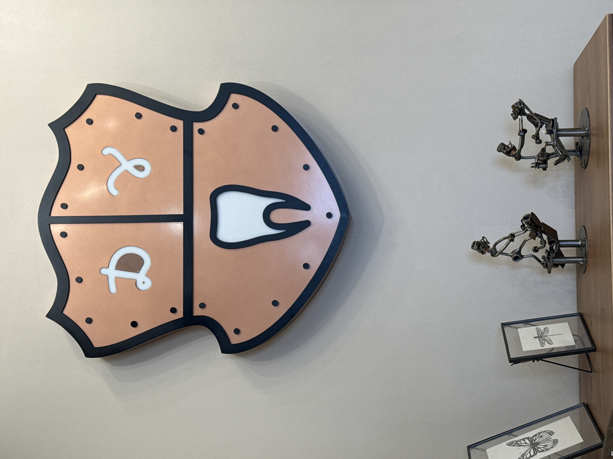 The image shows a dental office interior. A stylized, shield-shaped logo is mounted on a beige wall. The logo is divided into three sections. The top left section has a stylized Cyrillic letter resembling "Я". The bottom left section contains a stylized Cyrillic letter resembling "Д". The right section features a white, stylized tooth shape outlined in black. The shield shape has a black border with black circular elements spaced around the perimeter.
To the right of the logo, there are two metal sculptures resembling dentists operating on patients, mounted on a wooden shelf. Below the sculptures, there are two small framed prints with drawings of insects - a dragonfly and a butterfly.
The location is presumed to be a dental office in Izhevsk, Russia, based on the provided information.