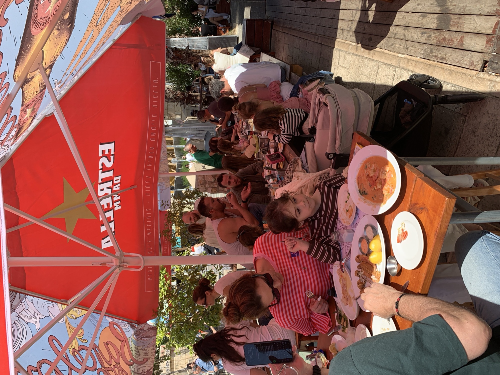 A family or a large group is seated at a long table, dining at an outdoor restaurant. A prominent red "Estrella Damm" branded sun umbrella offers shade. A variety of dishes, including what appears to be seafood, are on the table. Several people are engaged with their cell phones while others eat. The restaurant is located in a densely built area, likely in the city of Tel-Aviv, Israel. A stroller is parked at the edge of the table.