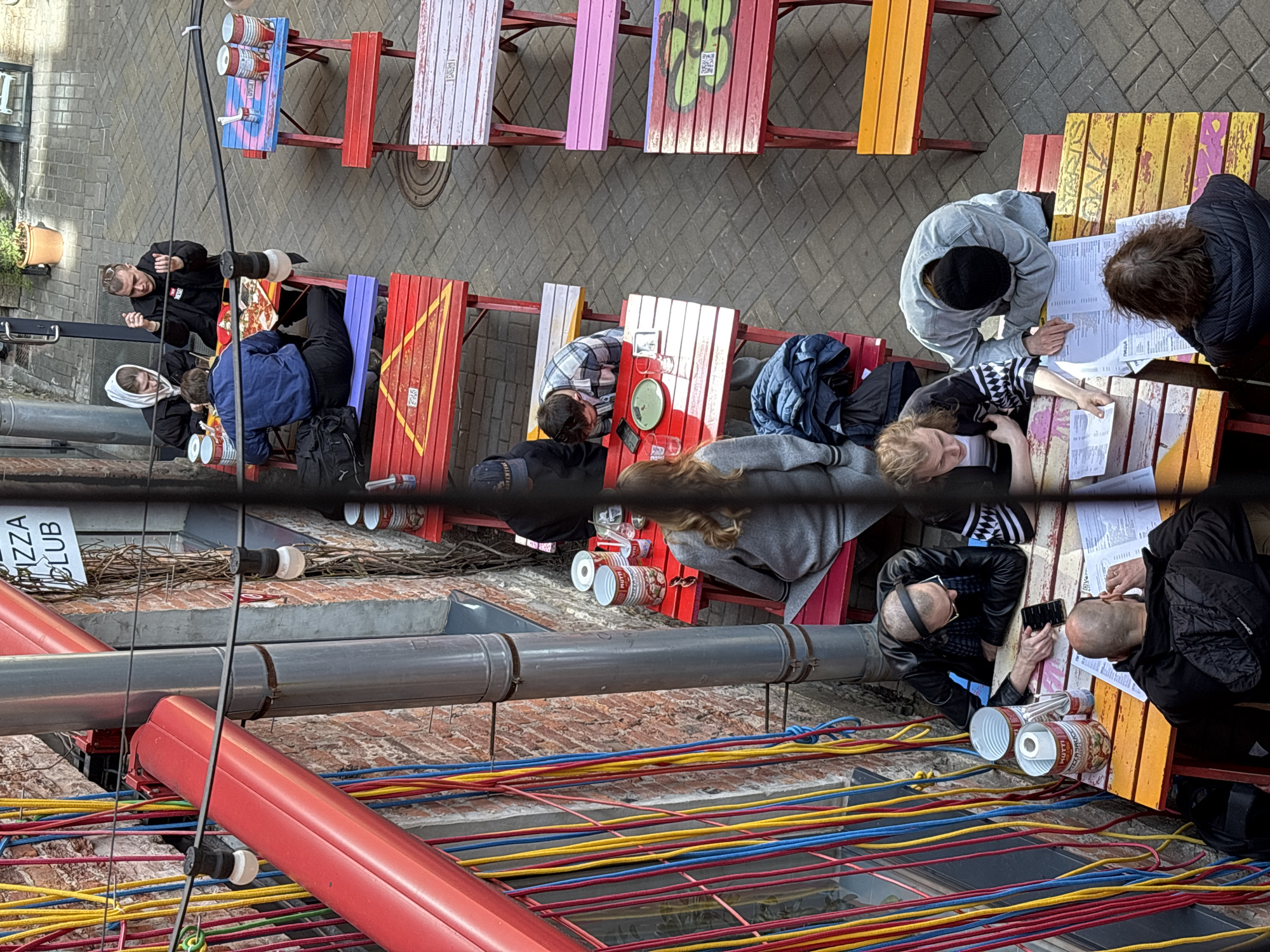 An eye-level shot depicts an open-air pizzeria from an elevated perspective. Several picnic-style tables and benches are arranged along a brick-paved outdoor dining area. The tables exhibit varied colors, including blue, red, pink, and yellow. Patrons are seated at several tables, engaged in various activities such as reading menus, conversing, and consuming food. In the foreground, a red structure with brightly colored horizontal wires - red, yellow, blue - forms a decorative element, possibly overhead. Above this, exposed pipes run along a brick wall, adding an industrial touch to the scene. A sign reading "PIZZA CLUB" is partially visible. The scene conveys an atmosphere of an urban eatery situated in Saint Petersburg, Russia.