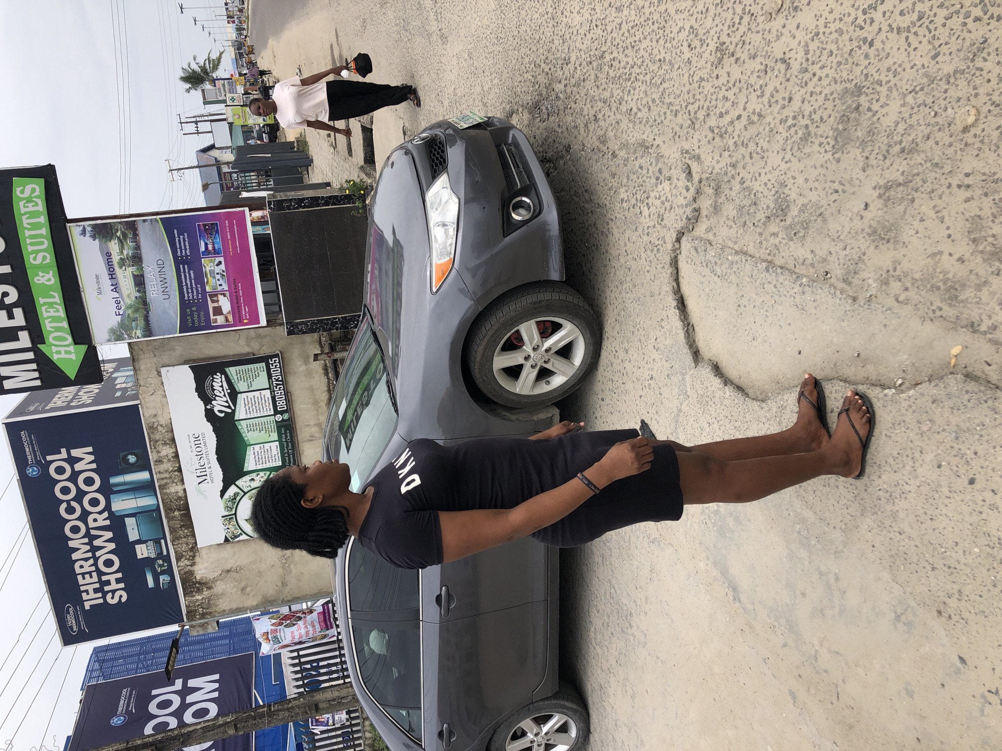 The image captures a scene in Ado, Nigeria. A woman in a black DKNY dress and flip-flops stands beside a gray car, looking up at several advertising signs. These include "THERMOCOOL SHOWROOM," "Milestone HOTEL & SUITES LIMITED" with restaurant menu items, and a "Milestone Hotel & Suites" advertisement featuring hotel amenities. Another man is seen walking in the background, wearing a light-colored shirt and dark pants, carrying a hat. The environment includes an urban street with cracked pavement.