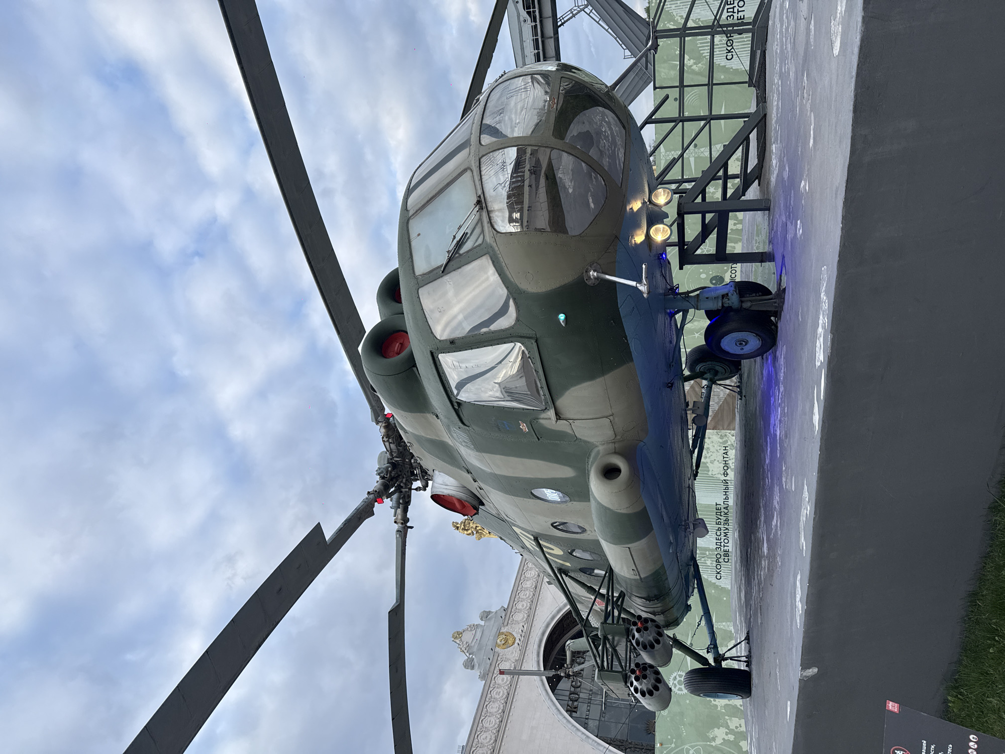 The image shows a military helicopter on display at the Exhibition of Achievements of National Economy (VDNKh) in Moscow, Russia. The helicopter is painted in a camouflage pattern of green and blue, with a large, multi-windowed cockpit and prominent rotor blades. It is mounted on a platform, and a portion of an ornate building or archway is visible in the background, adding to the location's unique atmosphere. The sky is overcast, with varying shades of gray. The display is positioned in an open-air setting, suggesting an outdoor exhibition area. The angle of the shot is from below, emphasizing the size of the helicopter. The display includes text, though the content is not readable.