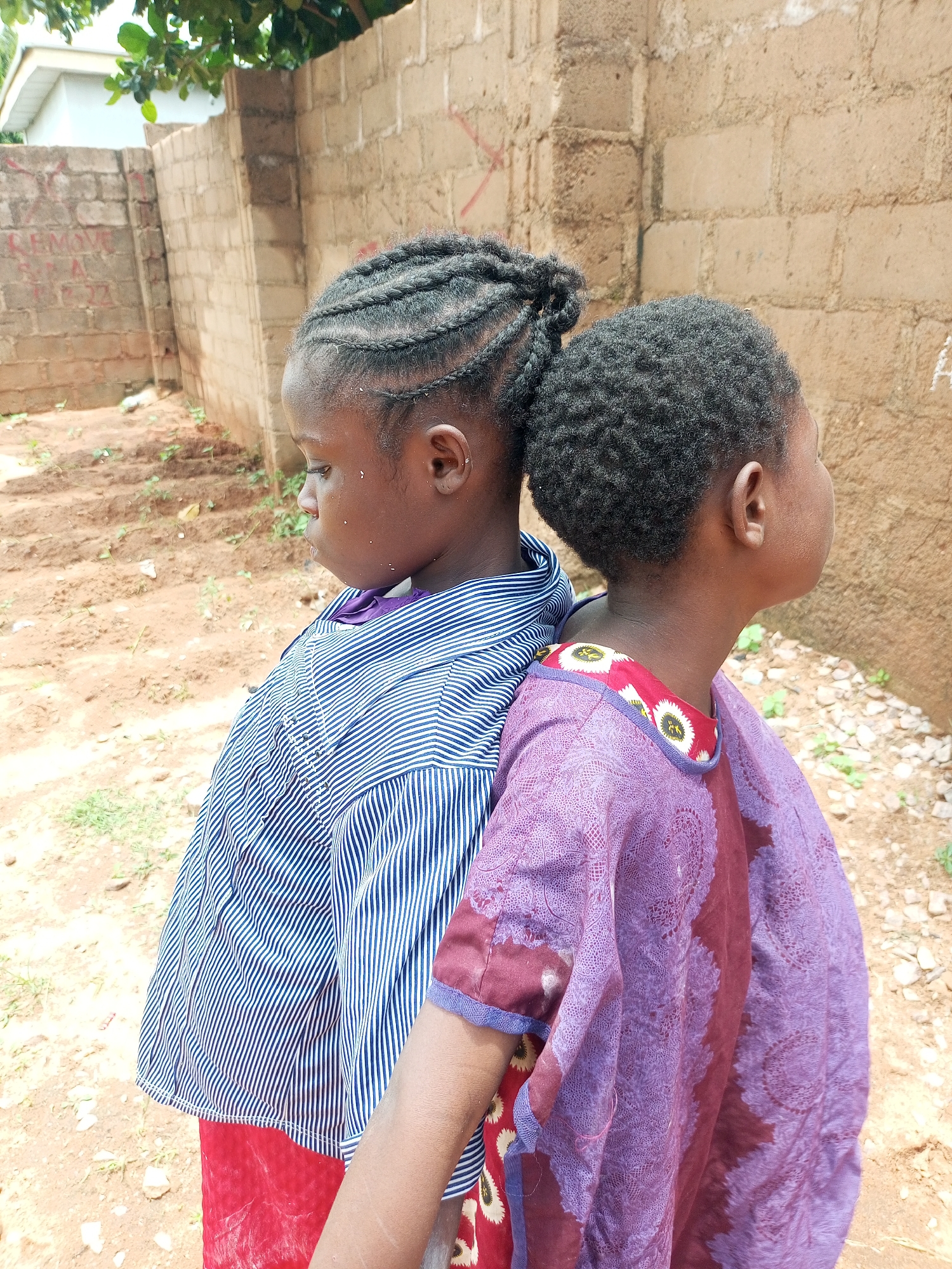 Two young girls stand back-to-back in an outdoor, dusty environment. The girl on the left is wearing a blue and white striped shirt and a red skirt, with her dark hair styled in braids pulled into a bun. The girl on the right has short, dark, curly hair and is wearing a purple garment with a patterned neckline. Behind them, a rough brick wall is visible, with some writing on it. The ground is dirt with sparse green vegetation and small rocks. The scene appears to be in Tempure, Nigeria.