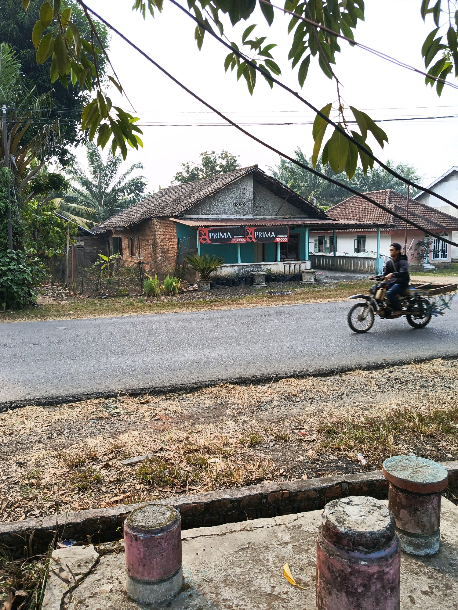 A man rides a motorbike with a flatbed trailer on a paved road in a rural area. The man is wearing a dark jacket and jeans, and the trailer appears to be carrying some sort of cargo. The road is lined with dry grass and some trees. In the background, there are several buildings, including a house with a red tiled roof and a smaller building with a banner that reads "PRIMA". The sky is bright and overcast. The scene appears to be in Wonosari, Indonesia.