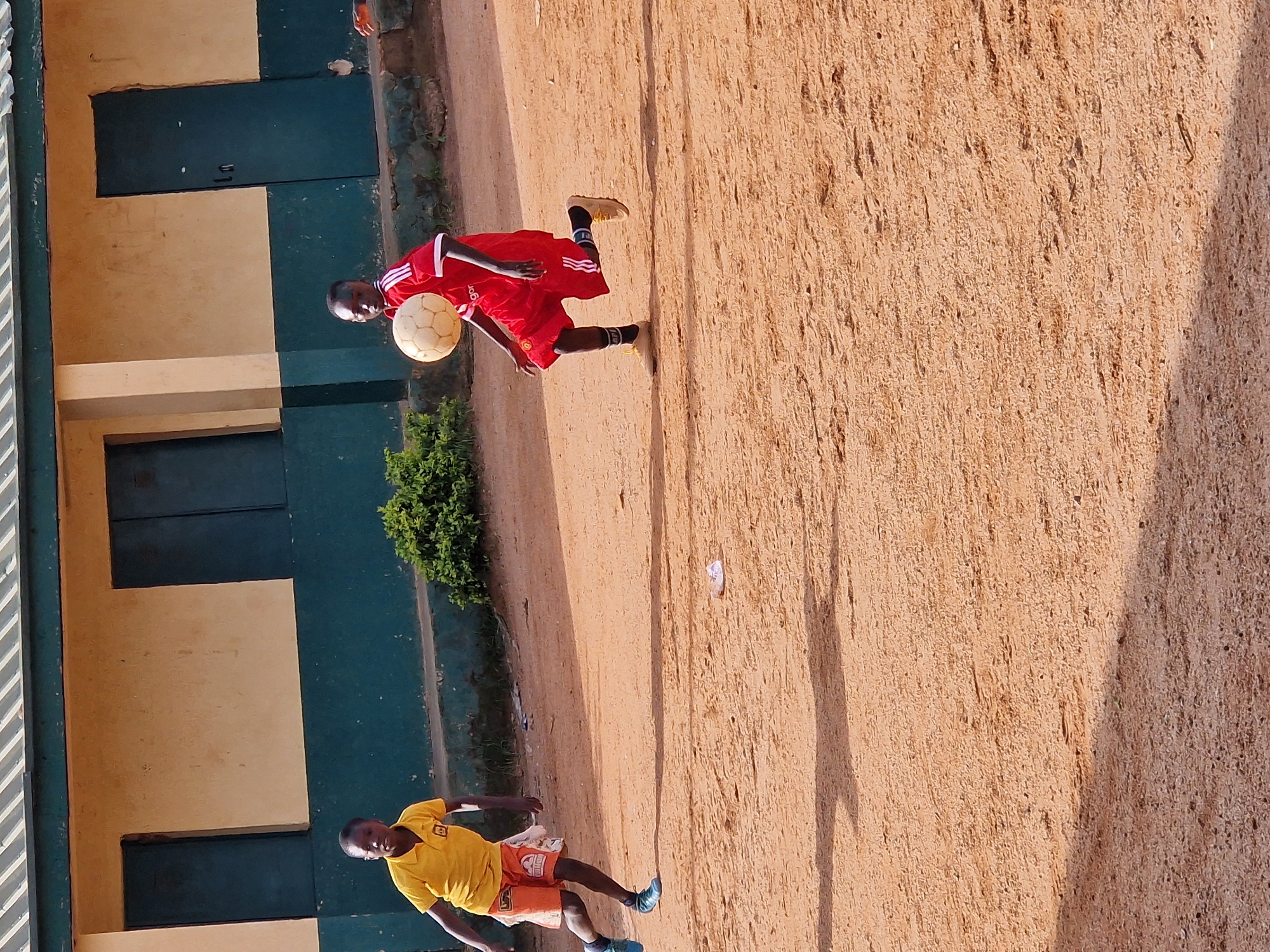 Two young boys are playing soccer on a dusty, outdoor field in front of a yellow and green building. The boy on the left is wearing a yellow shirt and orange shorts, and is running towards the right side of the frame. The boy on the right is wearing a red soccer jersey and shorts, and is dribbling a soccer ball with his head. The building appears to be a school, with large, dark-colored doors or windows visible. There are shadows on the ground, indicating it is daytime. The environment suggests an informal setting, possibly in Kabong, Nigeria, where children often play in open spaces.