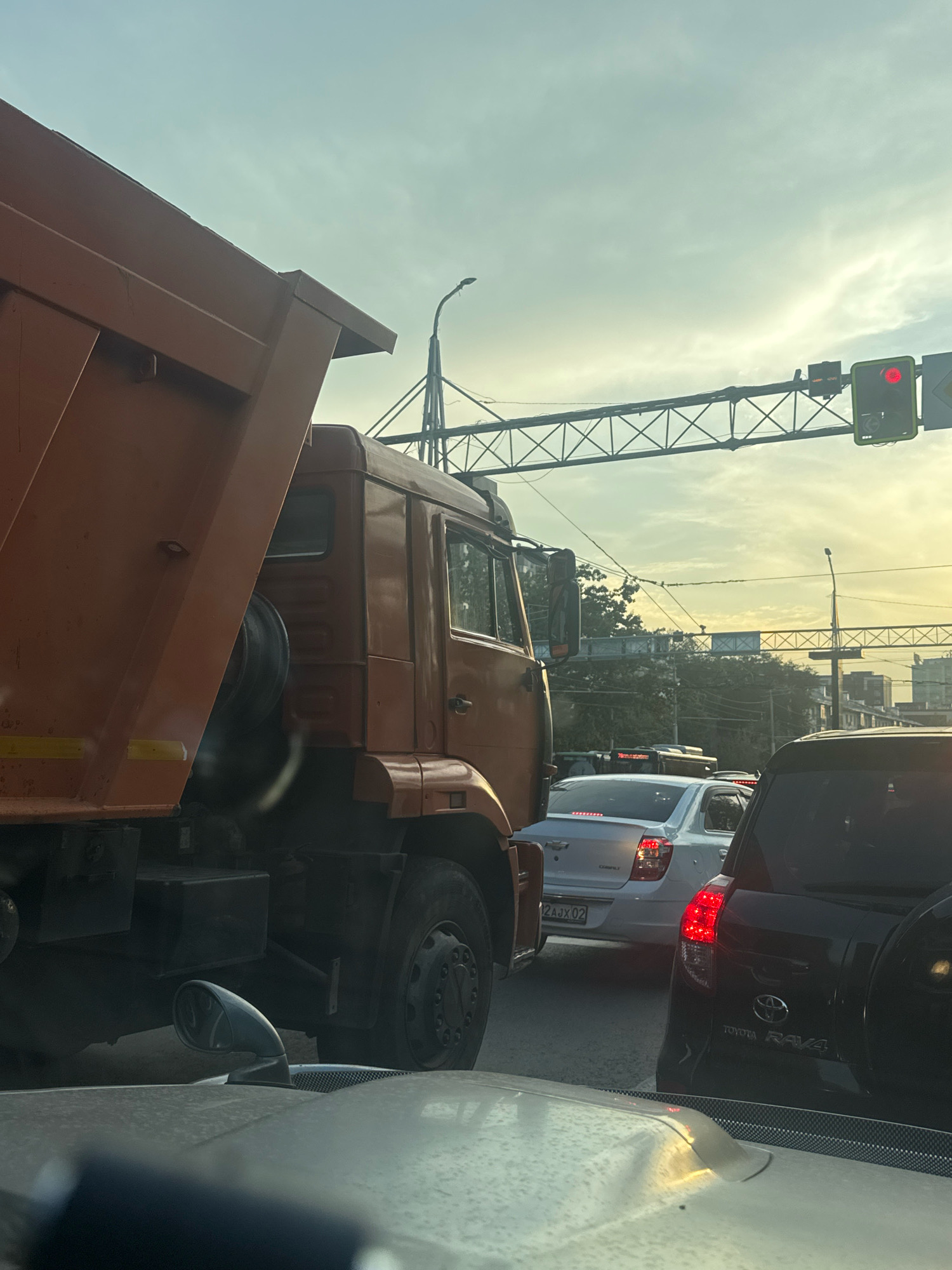 The image shows a traffic jam on a street in Almaty, Kazakhstan. The foreground features the hood of a car, with a side mirror of a large orange truck visible to the left. The truck appears to be a dump truck, with its bed raised. Behind the truck, several other vehicles are stopped in traffic, including a white sedan and a black SUV. The sky is overcast, with the sun setting in the distance, casting a warm glow. Traffic lights are visible overhead, indicating that the traffic may be stopped at an intersection. Buildings and trees line the street in the background. The overall scene depicts a common urban traffic scenario in Almaty.