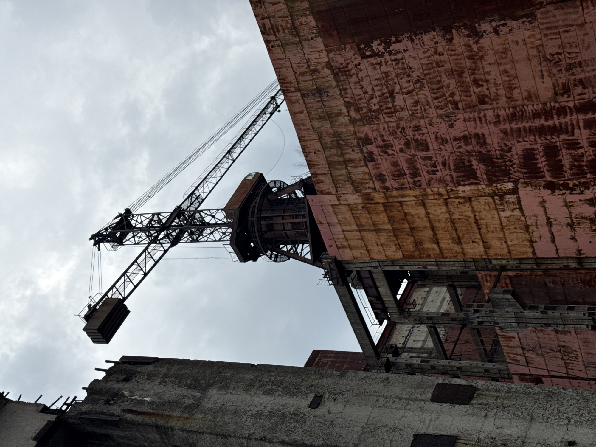 The image captures a scene of industrial decay, likely within the city of Pryp'yat', Ukraine, near the Chernobyl Nuclear Power Plant. An enormous crane, constructed of dark, intricate steel lattice, dominates the composition. The crane's lifting mechanism is suspended in the air.
Adjacent to the crane, a colossal, dilapidated building looms. The building's facade is a patchwork of rust-colored and faded pink panels, exhibiting severe corrosion and deterioration. Parts of the structure have collapsed.
In the foreground, crumbling concrete remnants suggest further wreckage and destruction. The sky is overcast. The scene evokes a sense of abandonment and the devastating impact of the Chernobyl disaster.