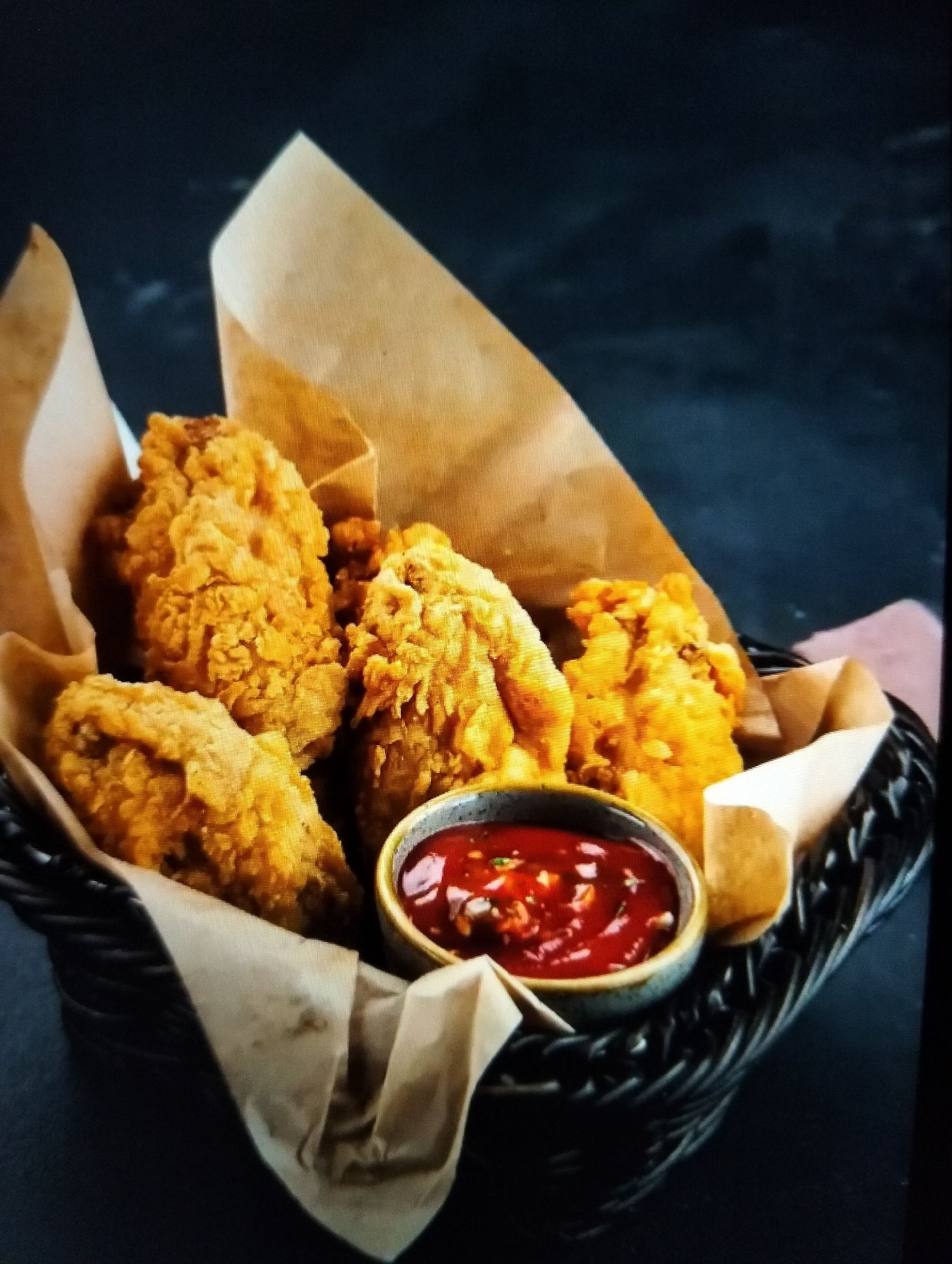 A close-up, eye-level, medium shot captures a woven basket filled with golden-brown, crispy fried chicken pieces, artfully arranged with brown parchment paper lining. A small, round, ceramic bowl of vibrant red sauce, likely a chili sauce or salsa, sits nestled among the chicken pieces. The background is a dark, textured surface, possibly a tabletop or wall, creating a moody and dramatic contrast that highlights the food. No people are visible, and the scene appears to be set for a food presentation or a restaurant setting. There is no discernible information about the location in Payagaboh, Indonesia.