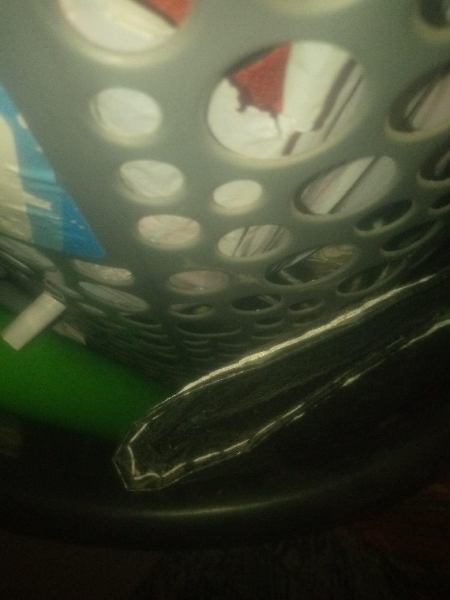 This is a low-angle, slightly blurry photograph of the interior of a laundry basket. The basket is made of gray plastic with a pattern of circular holes. Inside the basket, several items are visible, including what appears to be clothing. One item is red and white, possibly a towel or shirt. Another item is blue and white, possibly a plastic bag or packaging. The basket is resting on a dark surface, which could be the floor or another container. To the left of the basket, a bright green surface is visible, which could be a laundry mat or a plastic sheet. The lighting is dim, and there is a significant amount of noise in the image, making it difficult to discern finer details. This image was captured in Jauro Yinu, Nigeria.