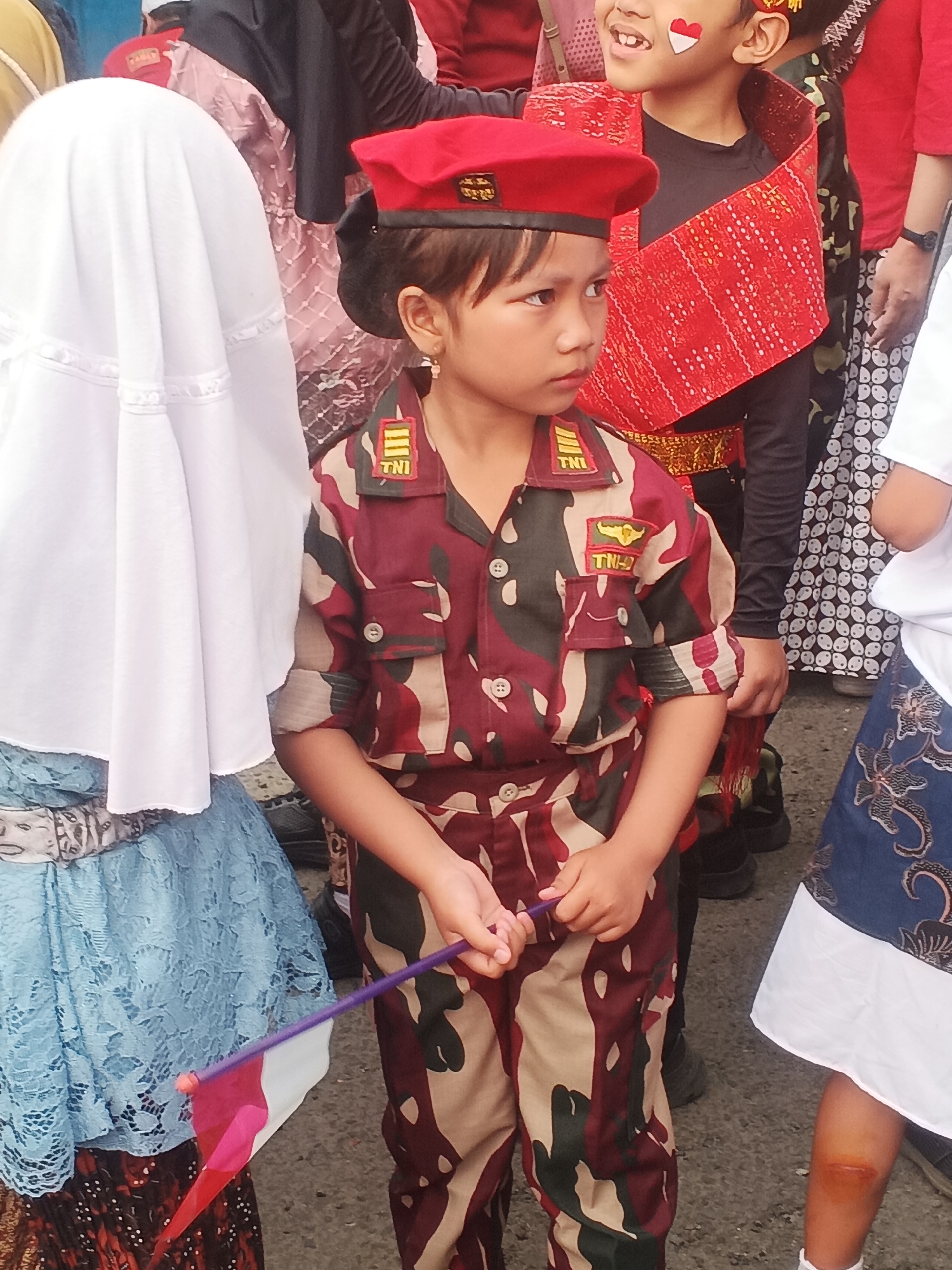 In the city of Sokanegara, Indonesia, a young girl, likely a child participant in a local event, is the central focus of the image. She is dressed in a camouflage military-style uniform, complete with a red beret and insignia bearing the letters "TNI". The uniform features a pattern of maroon, tan, and dark green. She is holding a small Indonesian flag, red and white, attached to a purple stick. Her left arm is bent, and her hand is in a relaxed grip on the flagstick, suggesting a moment of pause or transition.
To her left, a larger figure is partially visible, wearing a white hijab and a light blue lace dress, indicating the presence of other participants, possibly in traditional attire. To her right, another child is partially visible, wearing a black shirt with a vibrant red sash adorned with intricate patterns and gold trim. This child also has face paint, a red and white design resembling a heart on their cheek, symbolizing the Indonesian flag. Further to the right, another individual in a white shirt and a patterned skirt is present.
The background is indistinct, but suggests an outdoor setting, possibly a street or public area, with a blurred mixture of colors and shapes, typical of a crowd or a festive environment. The ground appears to be asphalt or a similar paved surface. The overall scene conveys a sense of cultural celebration or a children's event, with a strong patriotic theme, evidenced by the Indonesian flags and attire. The lighting suggests it is daytime, with the sun casting soft shadows.