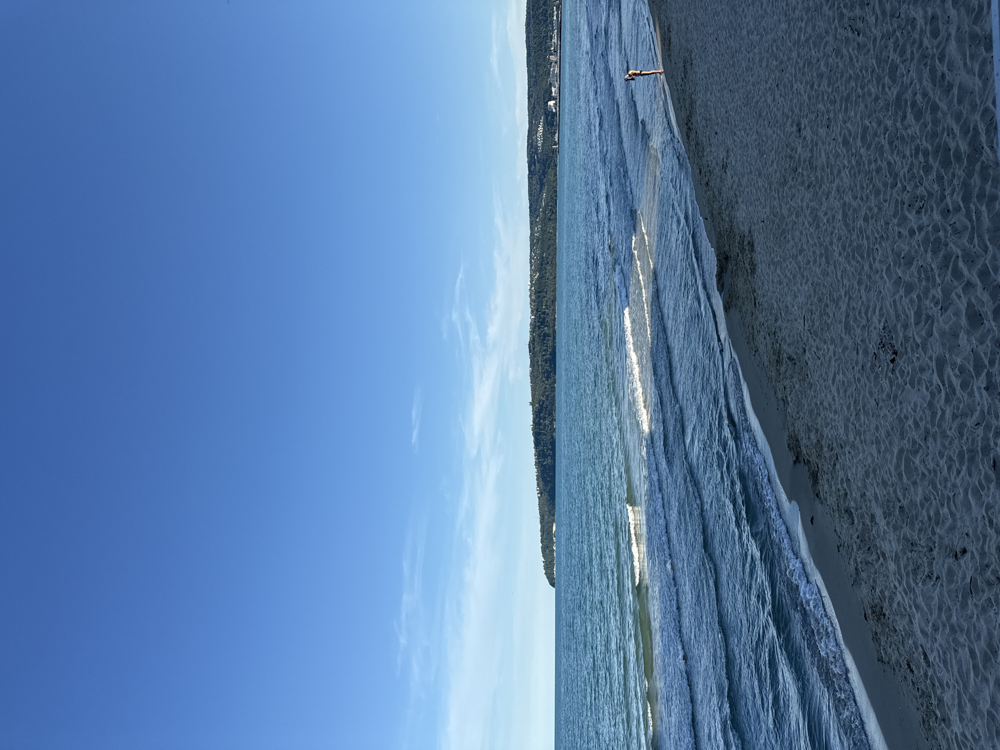 A wide shot of a beach on a clear, sunny day. The image is oriented vertically, with the sky occupying the left two-thirds and the ocean and beach on the right. The sky is a vibrant, cloudless blue. The ocean is a deep blue, with gentle waves rolling onto the shore. The beach is sandy and appears dry, with small ripples in the sand.
In the distance, along the shoreline, there is a line of trees and buildings. The trees are dense and green, and the buildings are scattered among them. The overall impression is one of a peaceful and serene beach scene.
A single person is visible in the water, standing near the shore. They are wearing a dark swimsuit and appear to be facing the ocean. Their posture is relaxed, and they seem to be enjoying the water.
The time of day appears to be daytime, likely late morning or early afternoon, given the brightness of the sunlight. The weather is clear and sunny, with no clouds visible in the sky. The location is a beach, likely a coastal area with a nearby town or resort.
The mood of the image is tranquil and inviting, conveying a sense of relaxation and enjoyment of the natural environment. There is no visible text in the image.