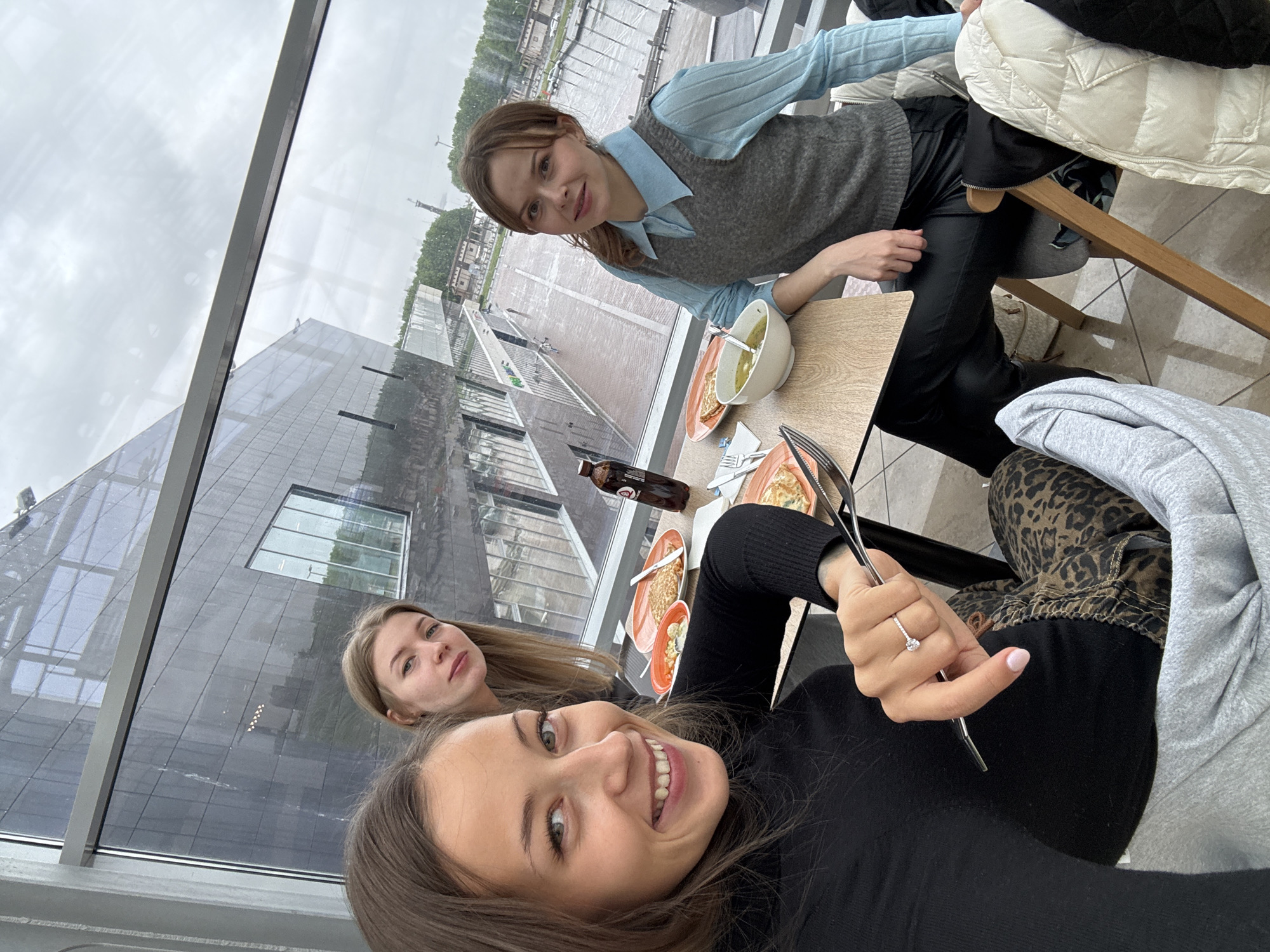 Three young women are seated at a table in an indoor location with a large window overlooking a cityscape. The woman closest to the camera is holding a pair of metal tongs and wearing a black long-sleeved shirt and a ring on her left ring finger. To her left and behind, another woman with blonde hair looks towards the camera. A third woman, wearing a light blue blouse with a gray sweater vest and black pants, is seated further back, also looking at the camera.
On the table, there are three orange plates, one white bowl, a bottle of Coca-Cola, and some cutlery. A building is visible outside, and, based on the architectural style and distant structures, the location appears to be in Saint Petersburg, Russia.
