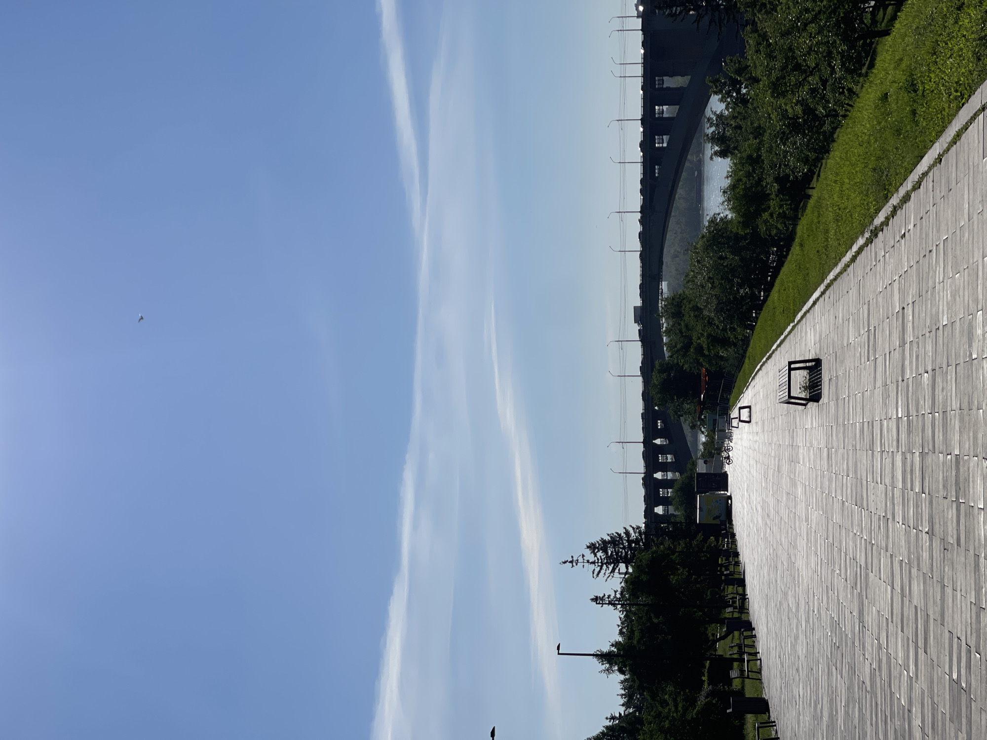 The image depicts a scenic view of the embankment in Krasnoyarsk, Russia, with the city's iconic arched bridge spanning the Yenisei River in the background. The foreground features a wide, paved walkway made of light-colored rectangular stones, creating a geometric pattern that leads the viewer's eye towards the bridge. Several dark-colored benches are strategically placed along the walkway, some empty and others occupied by silhouetted figures.
To the left of the walkway, a line of trees with lush green foliage creates a natural barrier, casting shadows on the path. A few streetlights are visible amongst the trees, suggesting the area is well-lit during the evening. On the right side of the walkway, a steep grassy embankment slopes down towards the river, adorned with more trees and bushes.
The Yenisei River itself is a shimmering expanse of blue water, reflecting the clear sky above. The bridge, a massive concrete structure with multiple arches, dominates the middle ground. Cars and trucks can be seen crossing the bridge, their headlights creating streaks of light against the dark concrete. The sky is a brilliant blue with a few wispy white clouds, and a lone bird is captured in mid-flight. The overall impression is one of a serene and picturesque urban landscape, ideal for a leisurely stroll or a moment of quiet contemplation.