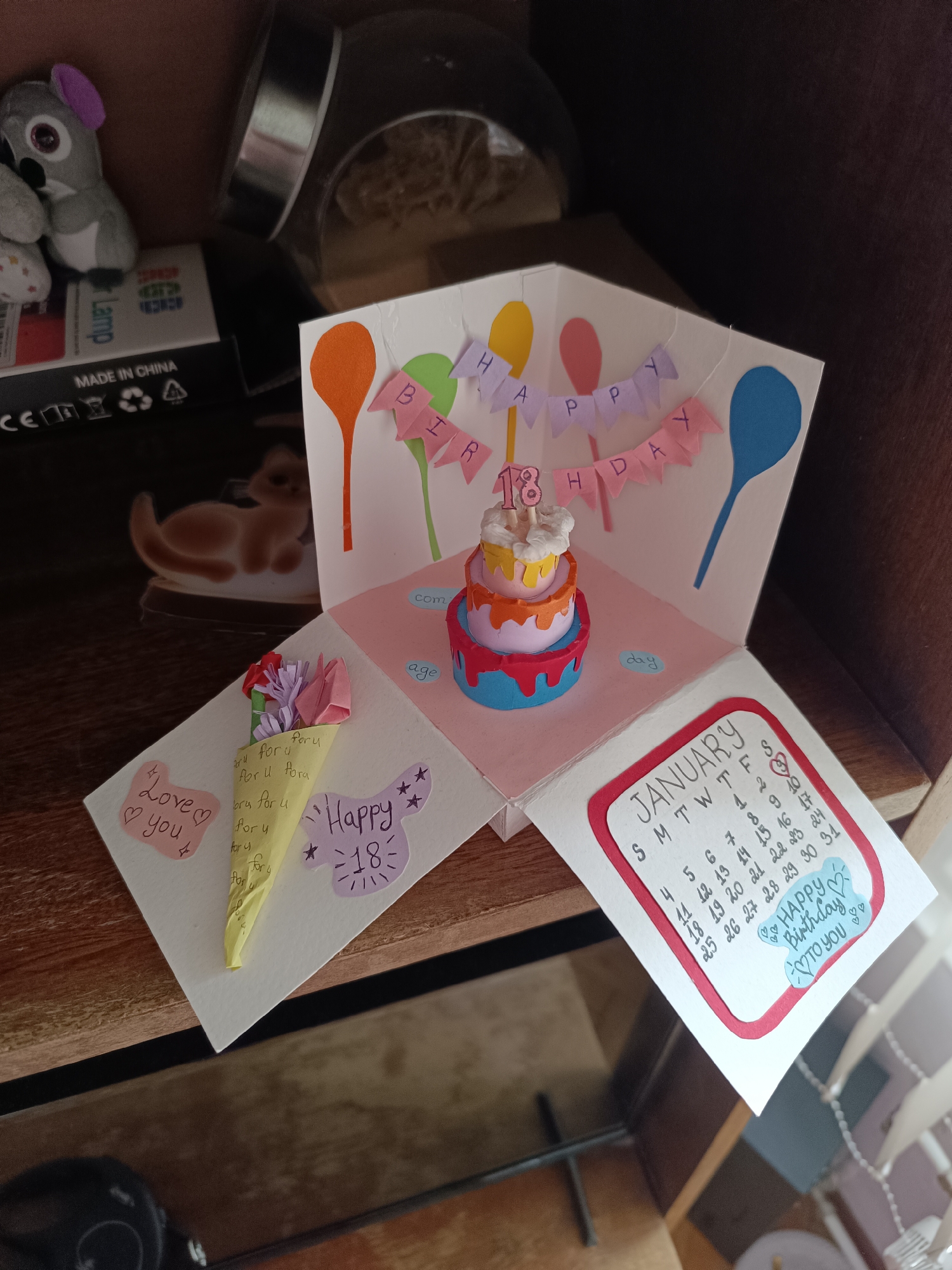 The image shows a handmade pop-up birthday card resting on a wooden surface. The card is open, revealing a miniature three-tiered cake with the number "18" on top, balloons, and a "Happy Birthday" banner. The card also features a bouquet of origami flowers in a yellow paper cone, a "Love you" note, a "Happy 18" message, and a January calendar with a heart drawn on the 16th and "Happy Birthday to you" written below.
Behind the card is a dimly lit area with a plush koala toy, a box labeled "Lamp," and a decorative cat figurine. A glass container is visible in the background. The setting appears to be indoors, possibly on a shelf or tabletop. The image was not taken in Pleven, Bulgaria.