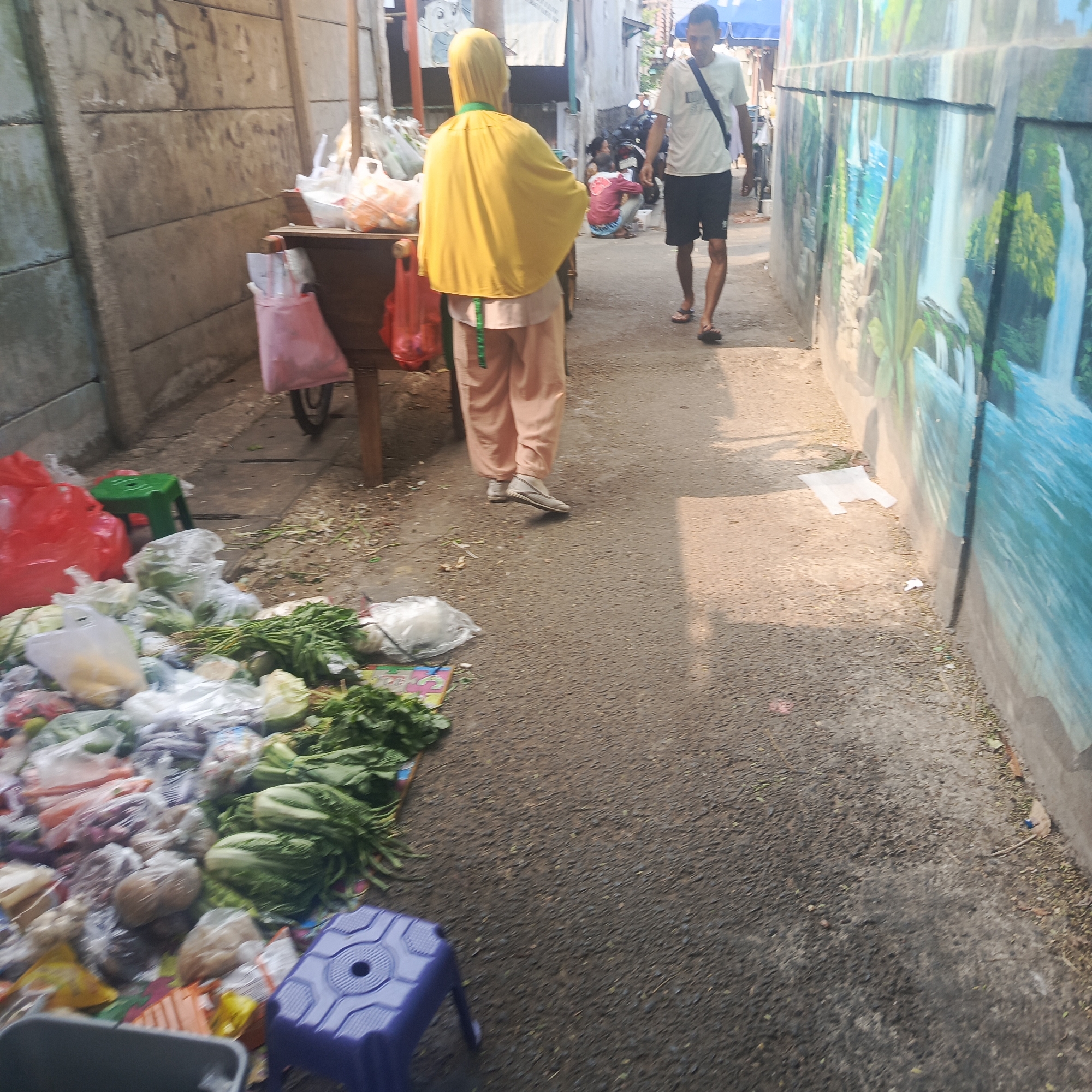 The image depicts a street market scene in South Jakarta, Indonesia. In the foreground, an assortment of fresh produce, including leafy greens and root vegetables, is displayed in plastic bags and on the ground. A small blue plastic stool is visible to the right of the produce. Further back, a person wearing a yellow head covering and light-colored pants stands near a wooden cart laden with items. To the right of the cart, a man in a white t-shirt and black shorts walks away from the camera. The background shows a mural of a waterfall on a wall, and various motorcycles and structures are present, suggesting a bustling urban environment. The lighting indicates it is daytime, with shadows cast across the ground.