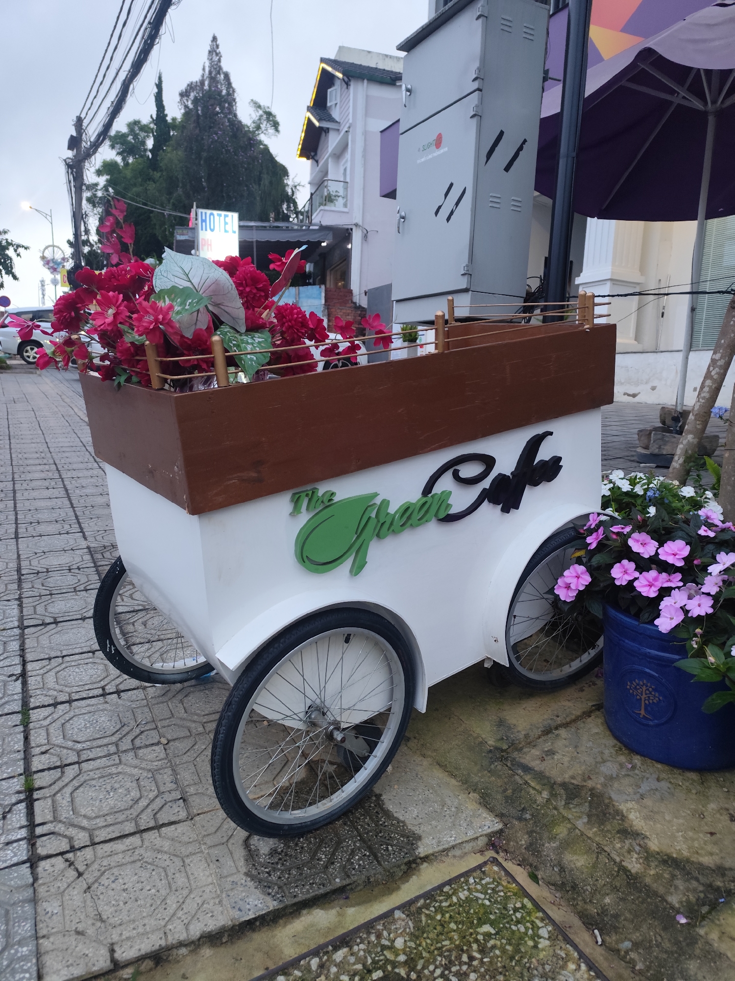 The image shows a white, three-wheeled cart with a wooden planter box filled with red artificial flowers and greenery. The cart has "The Green Cafe" written on its side in green and black lettering. It is parked on a tiled sidewalk in front of a building. To the right of the cart, a blue pot overflows with pink flowers. In the background, a tall building with lights is visible, along with trees and utility poles with many wires. The sky is overcast. This scene is typical of a street in Da Lat, Vietnam, likely a tourist area with cafes and shops.